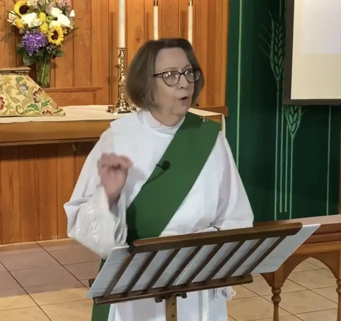 A woman dressed in a white robe with a green sash standing behind a wooden podium, gesturing with her right hand, with a wood-paneled background, flowers, and candles behind her.