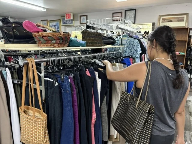 Woman shopping for pants at a thrift store, browsing a rack filled with various colored pants and skirts, with handbags and photo frames on the shelves above.