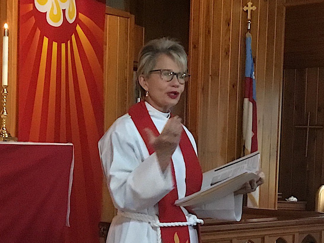 A woman dressed in a white robe with a red scarf and rope belt, standing at a pulpit in a church, holding a paper and speaking. Behind her are wooden walls, a flag, and a large red and yellow banner with religious symbols.