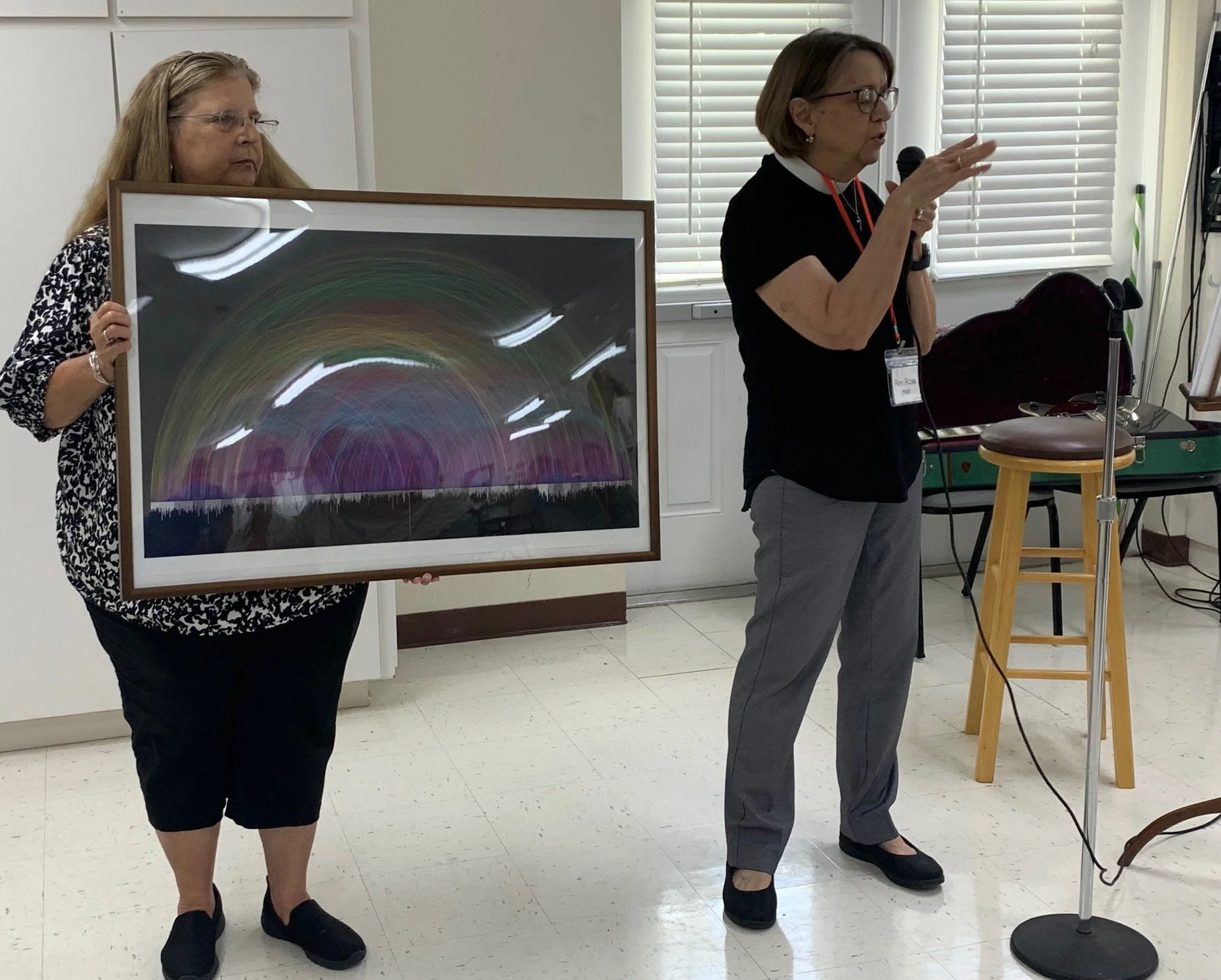 Two women, one holding a framed artwork and the other speaking into a microphone, in a room with office furniture and windows with blinds.