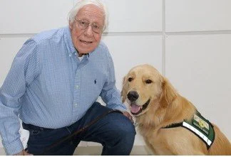 Older man with white hair and glasses smiling next to a golden retriever wearing a service vest.