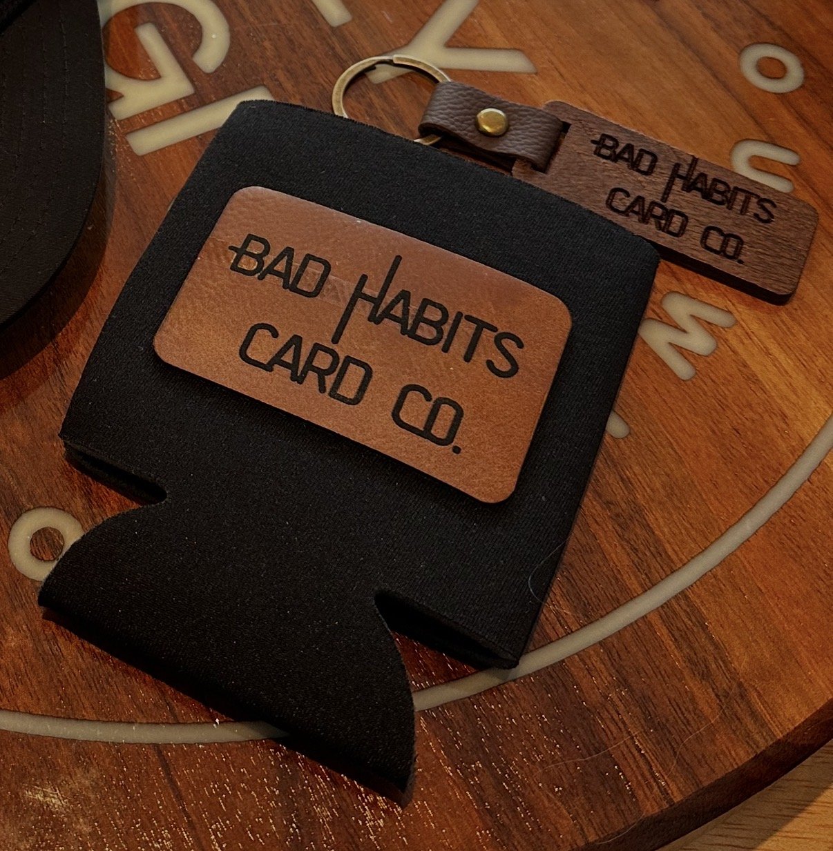 A black cardholder with a brown label that reads "Bad Habits Card Co." and a separate brown leather keychain with the same label, placed on a wooden surface with scattered beige plastic numbers around them.
