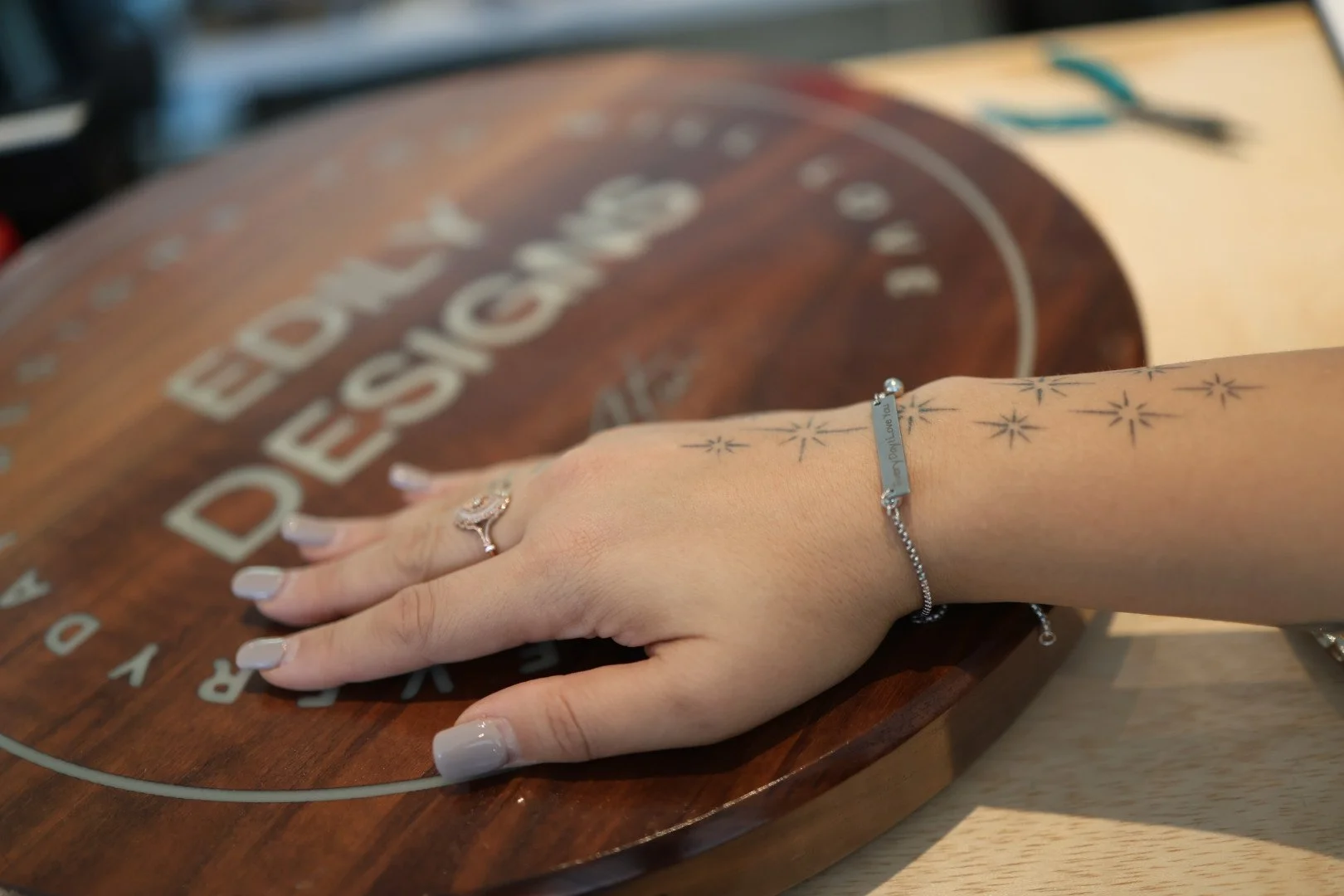 A person's left hand resting on a round wooden surface with the words 'Red Dog Salvage' partially visible. The person is wearing a silver ring with a gemstone on their finger, a silver bracelet, and has star-shaped tattoos on their wrist and forearm. The background is slightly blurred.