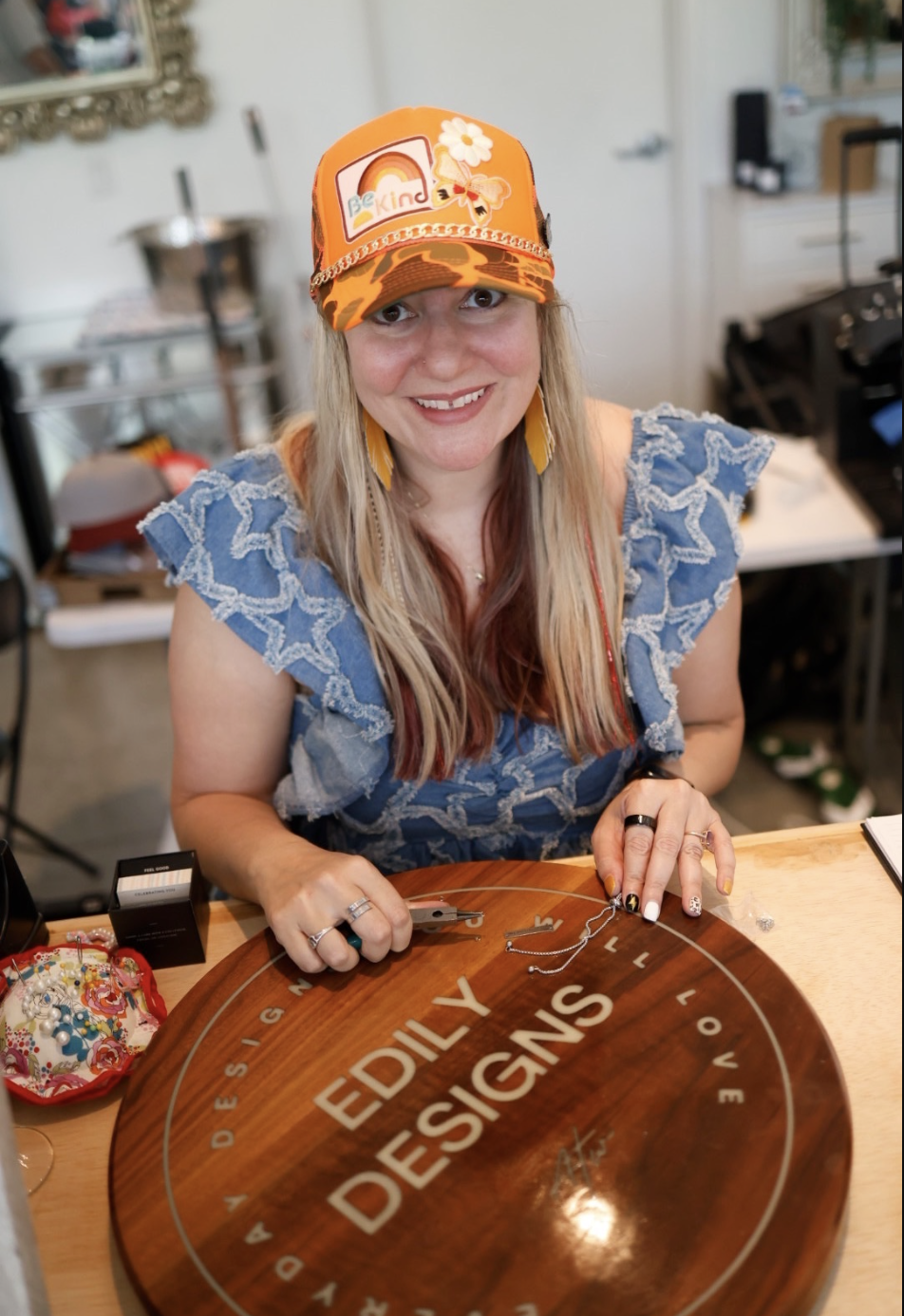 A smiling woman with long blonde hair, wearing a camouflage orange cap with floral and butterfly patches, sits at a table with jewelry tools, a blue ruffled blouse, and a circular wooden sign for Edily Designs.