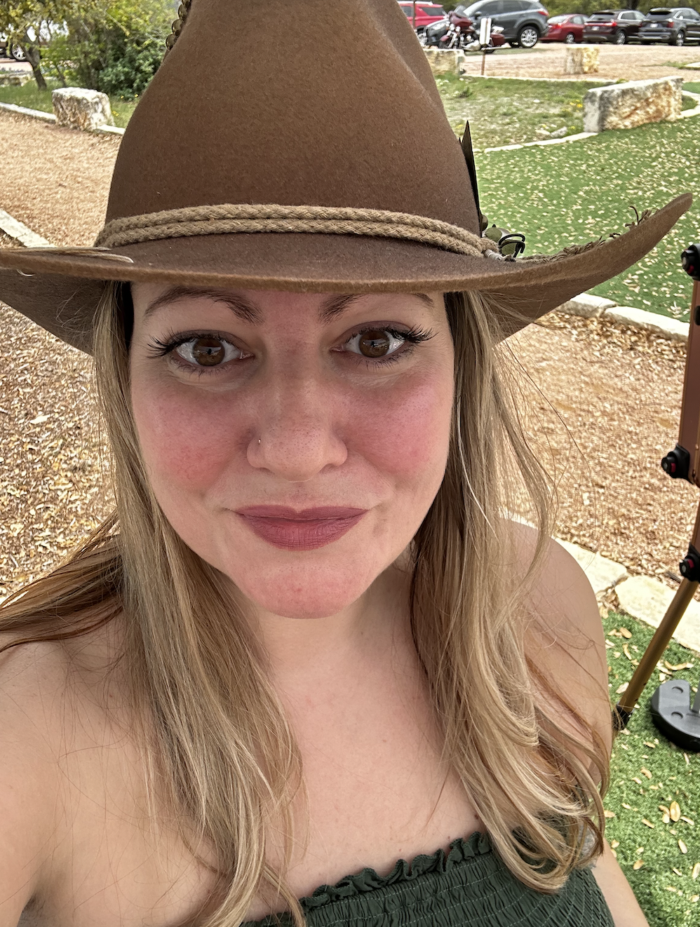 A woman with long blonde hair wearing a brown cowboy hat with a rope band, smiling outdoors in a park with trees, rocks, and parked cars in the background.