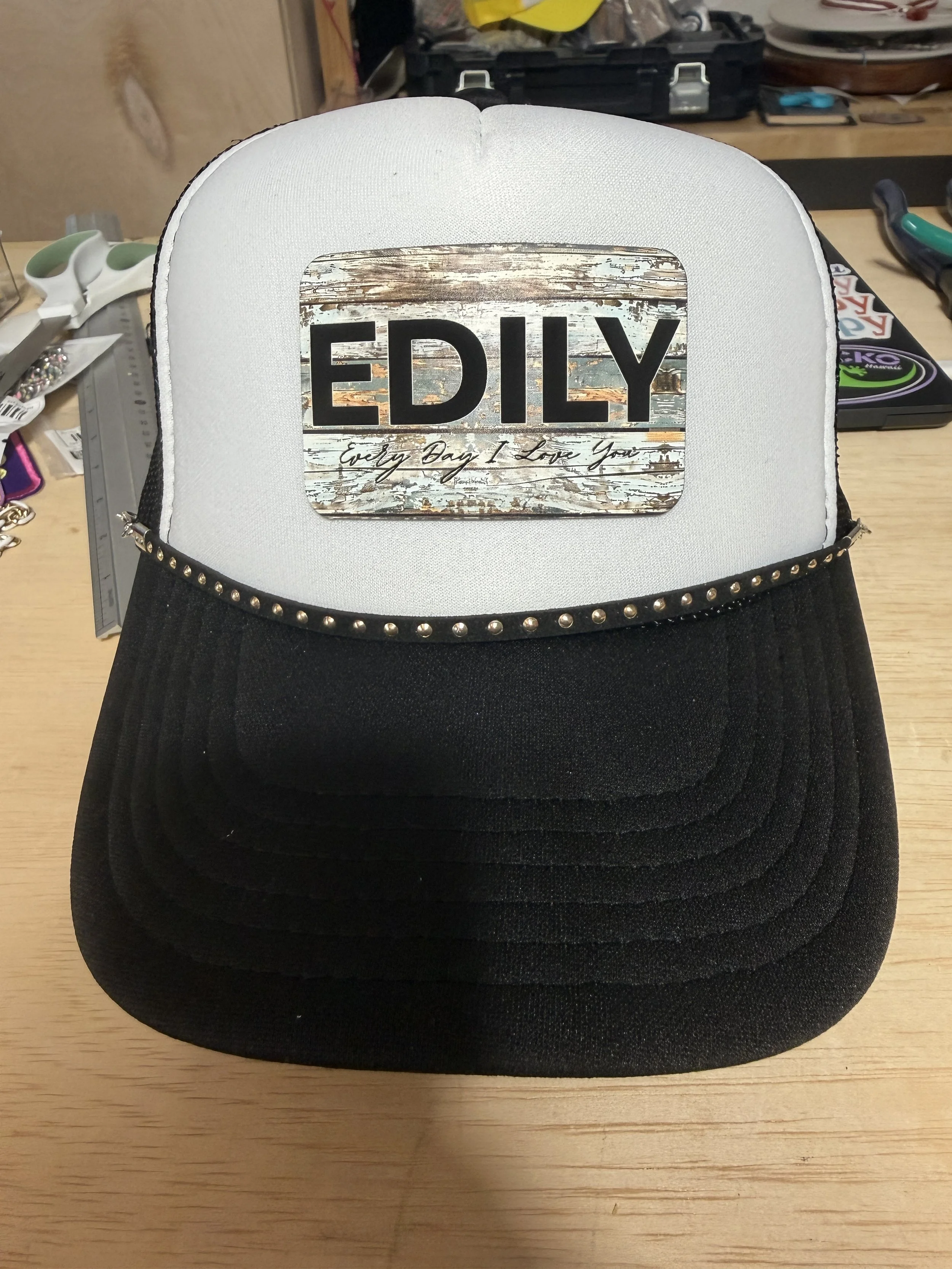 A white and black trucker hat with a rectangular patch on the front that says 'EDLY' and 'Every Day I Love You' beneath it, placed on a wooden surface.