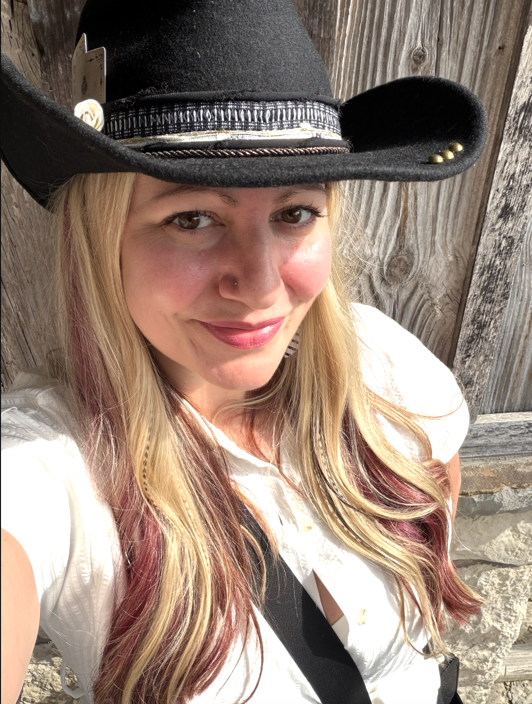 A woman taking a selfie outdoors, wearing a black cowboy hat, with long blonde and reddish hair, standing against a wooden fence.