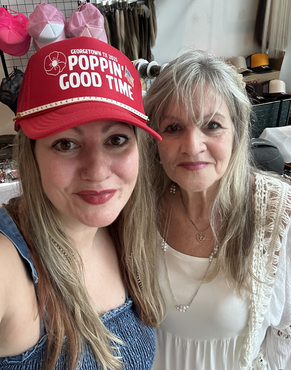 Two women taking a selfie indoors. The woman on the left is wearing a red cap with the text 'Poppin' Good Time' and a blue top. The woman on the right is wearing a white top and a beige crochet cardigan with jewelry.