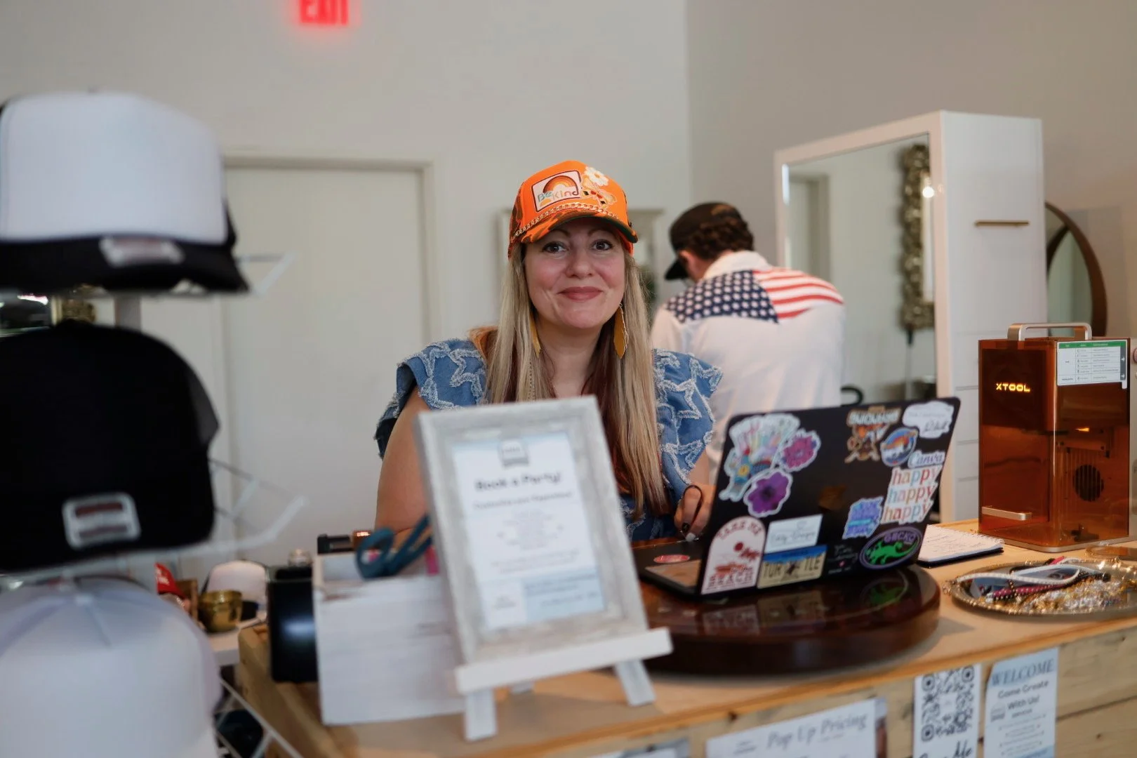 A woman with long blonde hair wearing an orange cap and colorful earrings smiling at a table with various items including a laptop covered in stickers, a framed sign, and merchandise. In the background, a person in a shirt with an American flag pattern is facing away from the camera.
