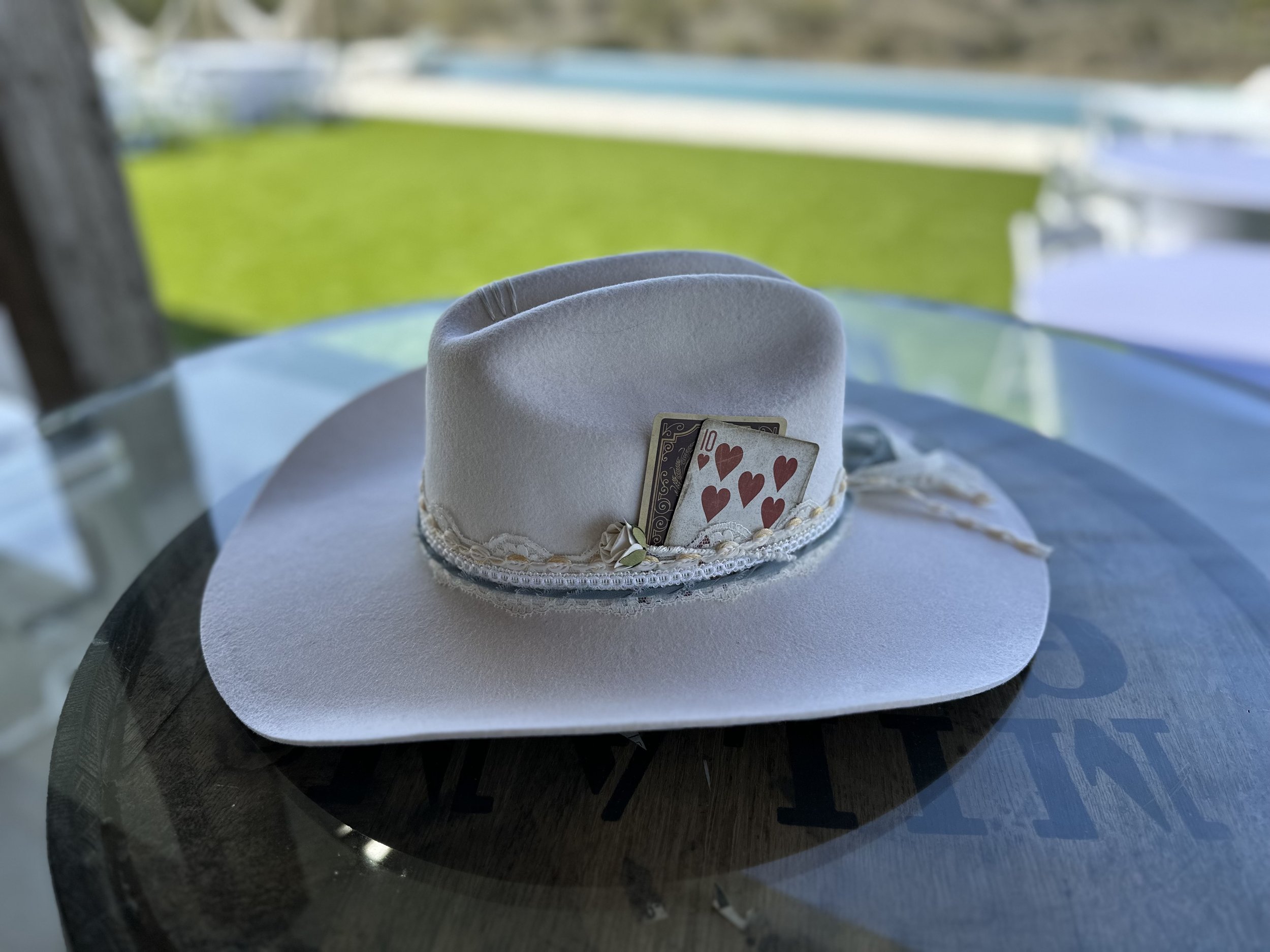 A white hat with a decorative band and playing cards tucked into the band, placed on a round table with a blurred green outdoor background.
