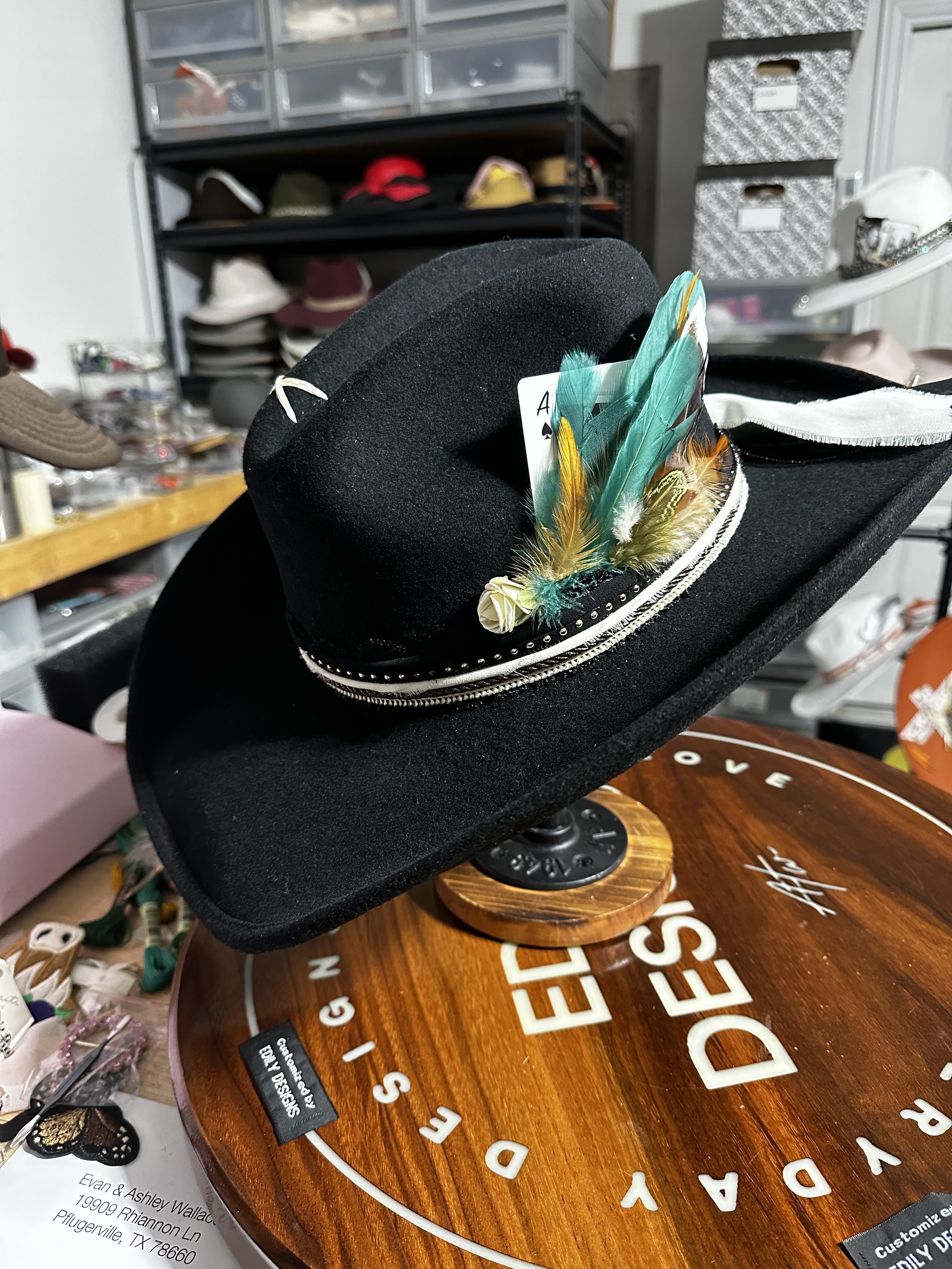 Close-up of a black felt hat with colorful feathers, a white playing card, and decorative trim, displayed on a wooden surface with large white letters.