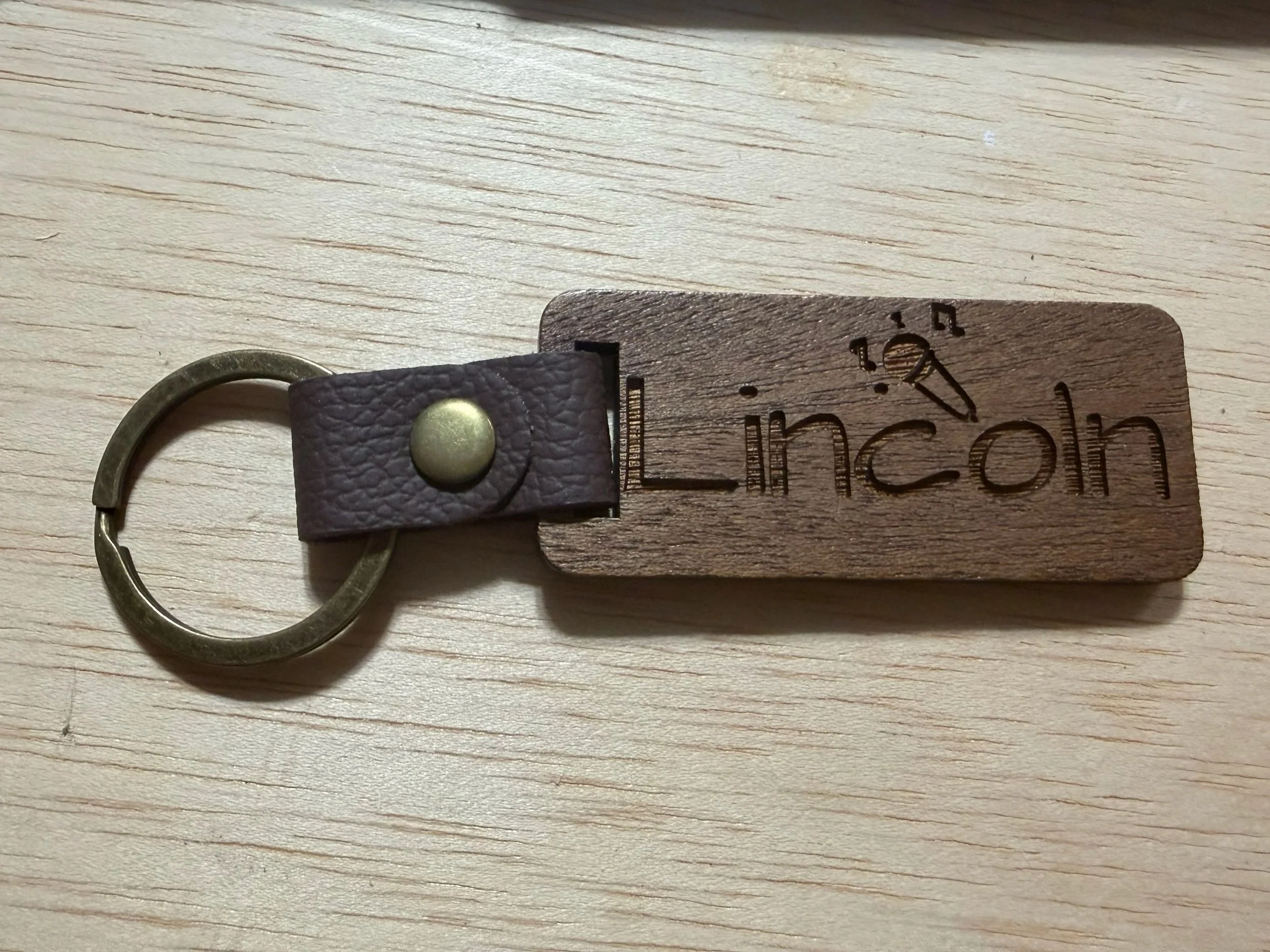 A close-up of a keychain with a brown leather strap and a metal keyring resting on a light wooden surface. The leather strap has engraved text and symbols.