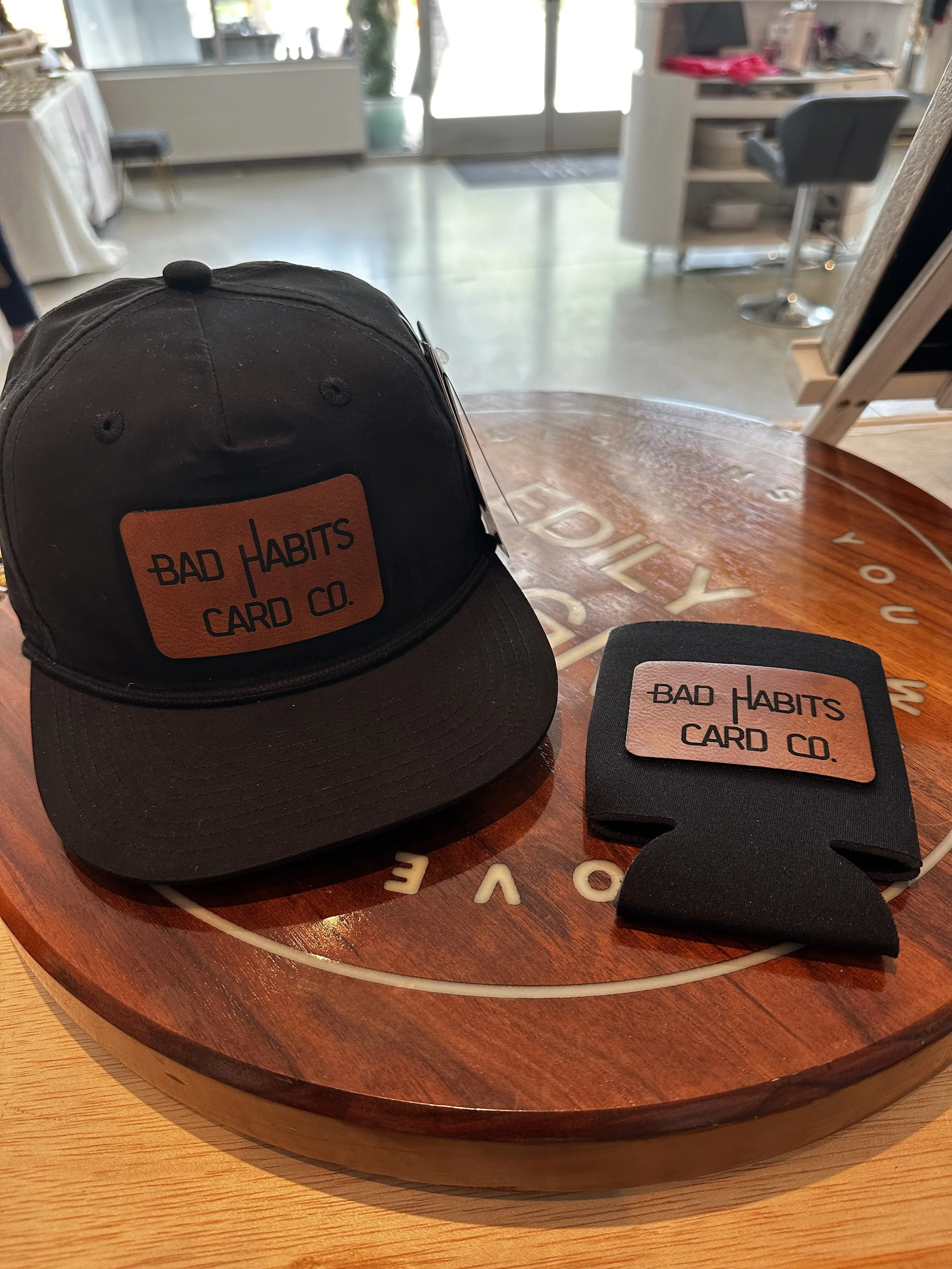 A black baseball cap and a black cardholder with orange and black labels, both labeled 'Bad Habits Card Co.', are placed on a round wooden table with a sign or logo underneath. The background shows a softly lit interior space with furniture and a door leading outside.