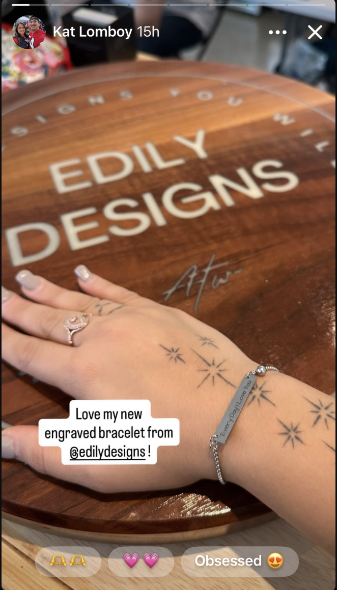 A hand with a bracelet, ring, and tattoos resting on a round wooden surface with the inscription 'EDILY DESIGNS' and the signature 'Art'.