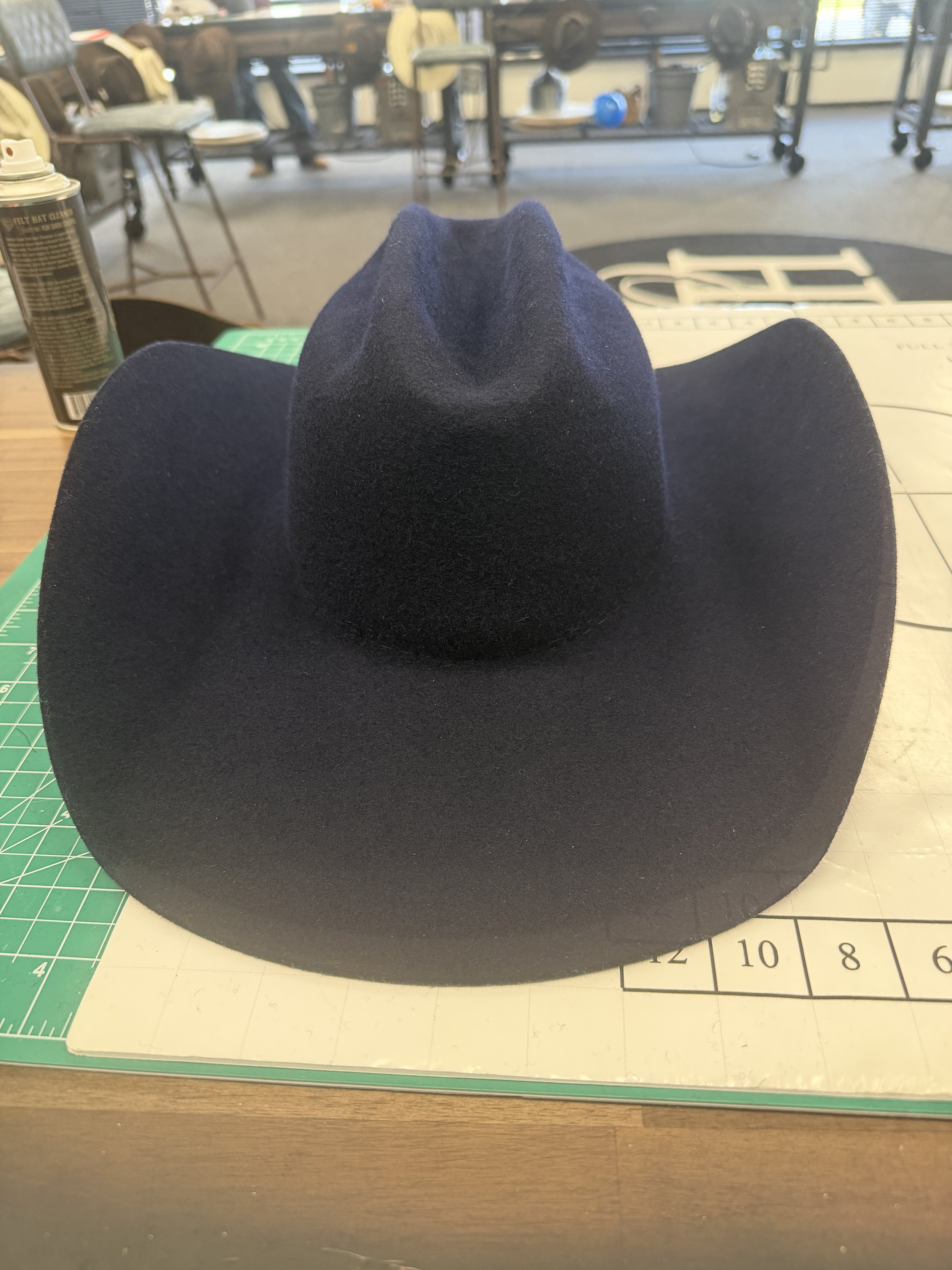 A black felt cowboy hat resting on a cutting mat on a wooden table, with a workspace in the background.