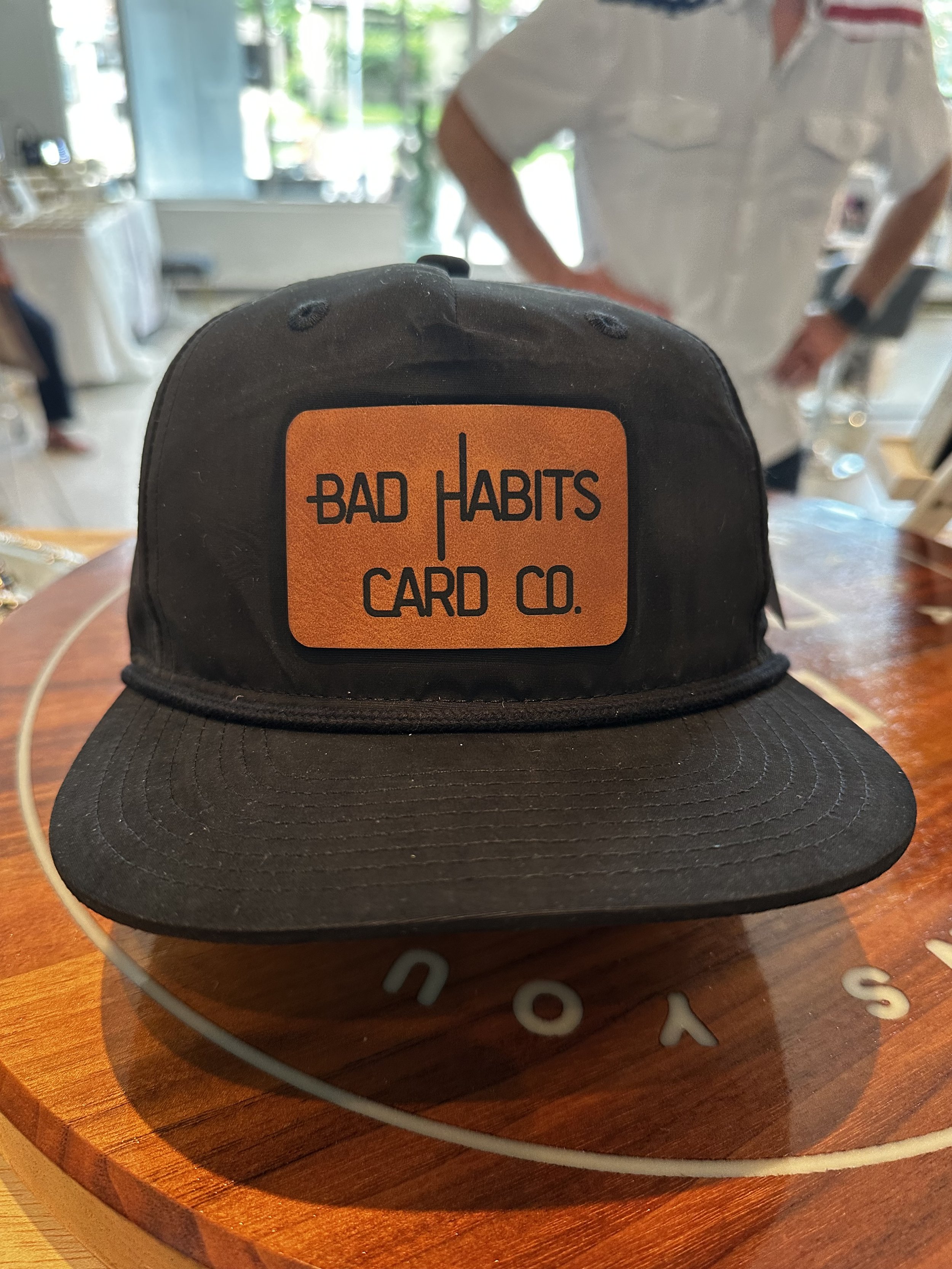 A black trucker cap with an orange patch that reads 'Bad Habits Card Co.'