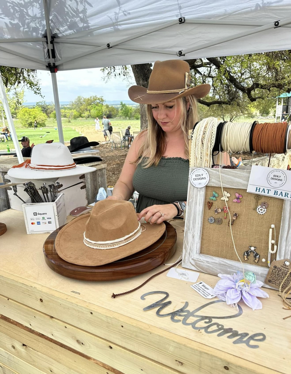 A woman at a hat bar shop, wearing a brown cowboy hat and a dark green top, displays a light brown hat with a white ribbon. The shop has various hats, jewelry, and accessories, with a "welcome" sign on the front of the rustic wooden counter. There are trees and people in the background under a white canopy.