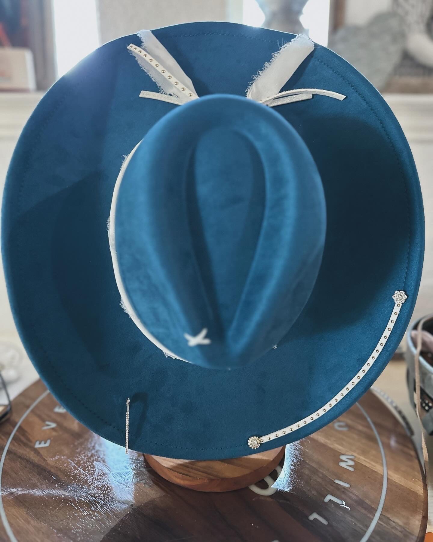 A blue cowboy hat with white decorative accents and rhinestones, placed on a wooden display stand on a table with a wooden surface.