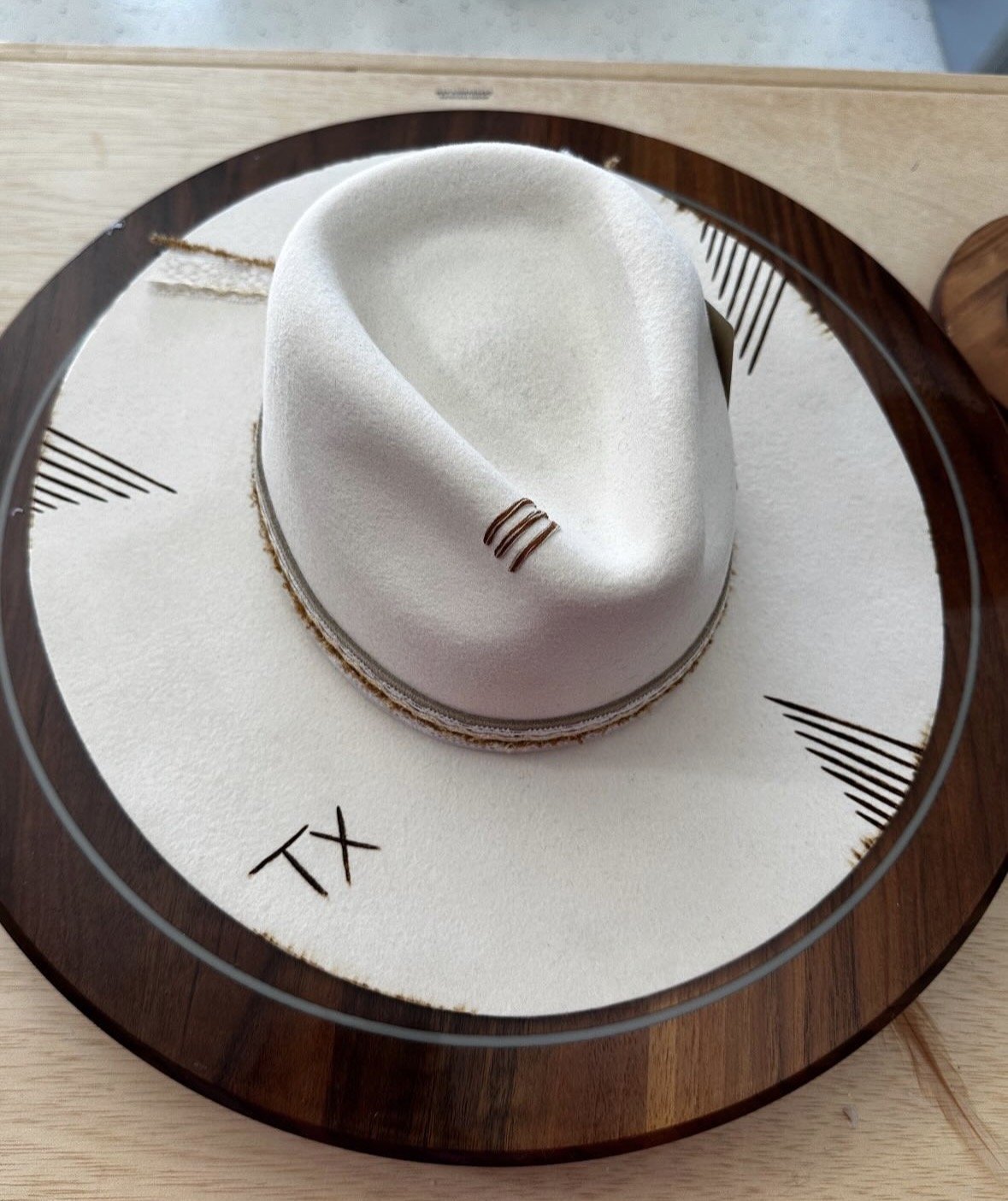 A white fedora hat with a brown band, placed on a white circular surface inside a glass top, with the initials 'TX' written on the surface.