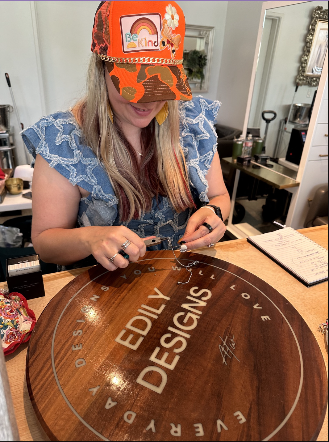 A woman wearing a colorful cap with a rainbow and the words "Be Kind" is working on jewelry at a desk. She is using tools to attach a chain to a a large wooden circular sign with the words "EDISTO ISLAND" engraved on it.