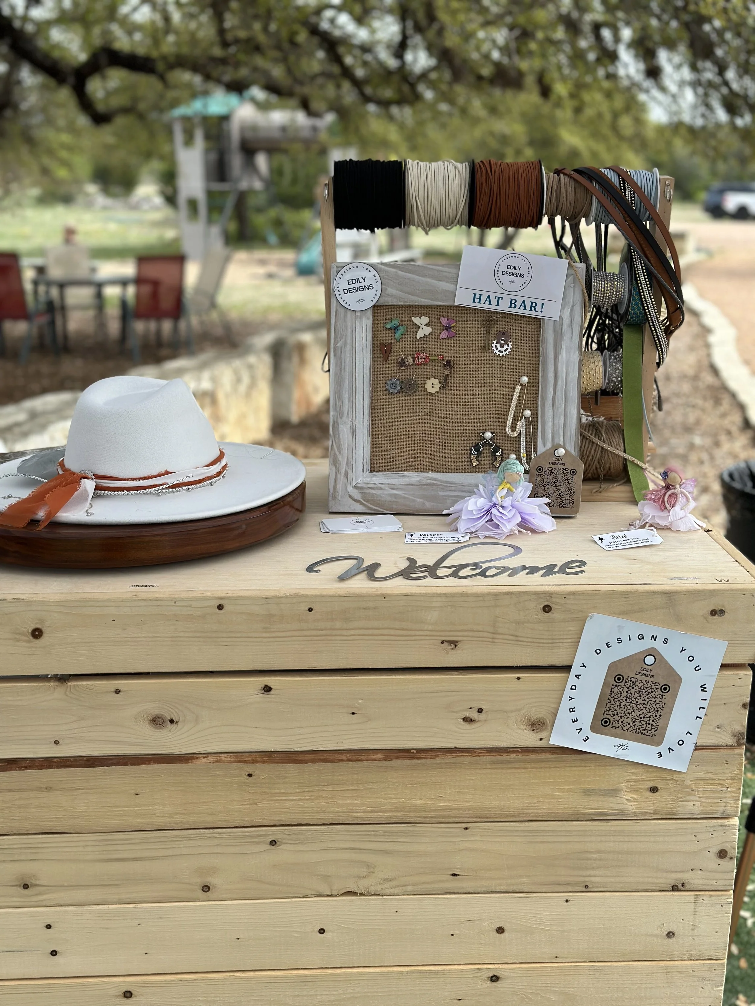 Wooden display stand with assorted jewelry and accessories, including bracelets, earrings, and a white hat with an orange ribbon, set outdoors near trees and a playground.