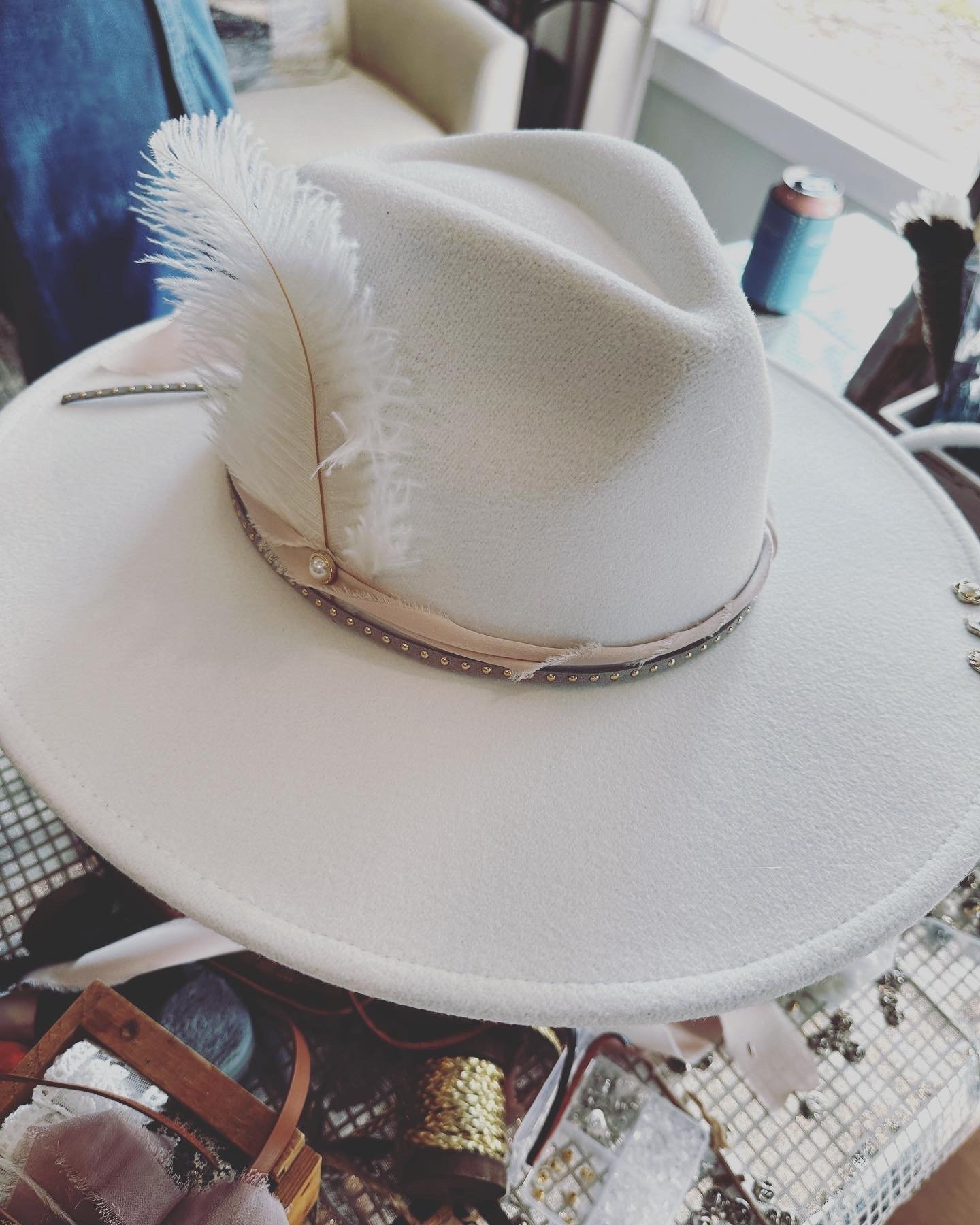 A white wide-brimmed hat with a feather and pearl embellishment on the band, resting on a surface near a window.