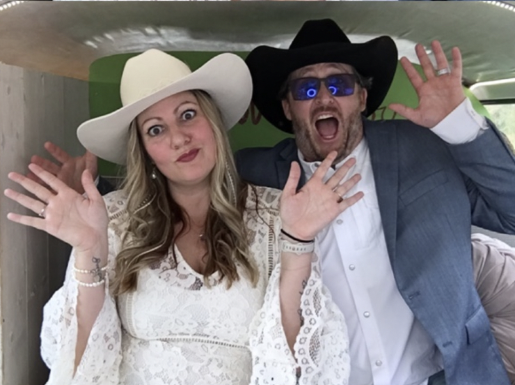 A woman and a man dressed in Western-style outfits, both wearing large cowboy hats, with the woman in a white hat and the man in a black hat. They are smiling and waving at the camera, with the woman's hand near her face and the man's hand raised in a greeting. The woman has long blonde hair and the man is wearing sunglasses with blue reflections.