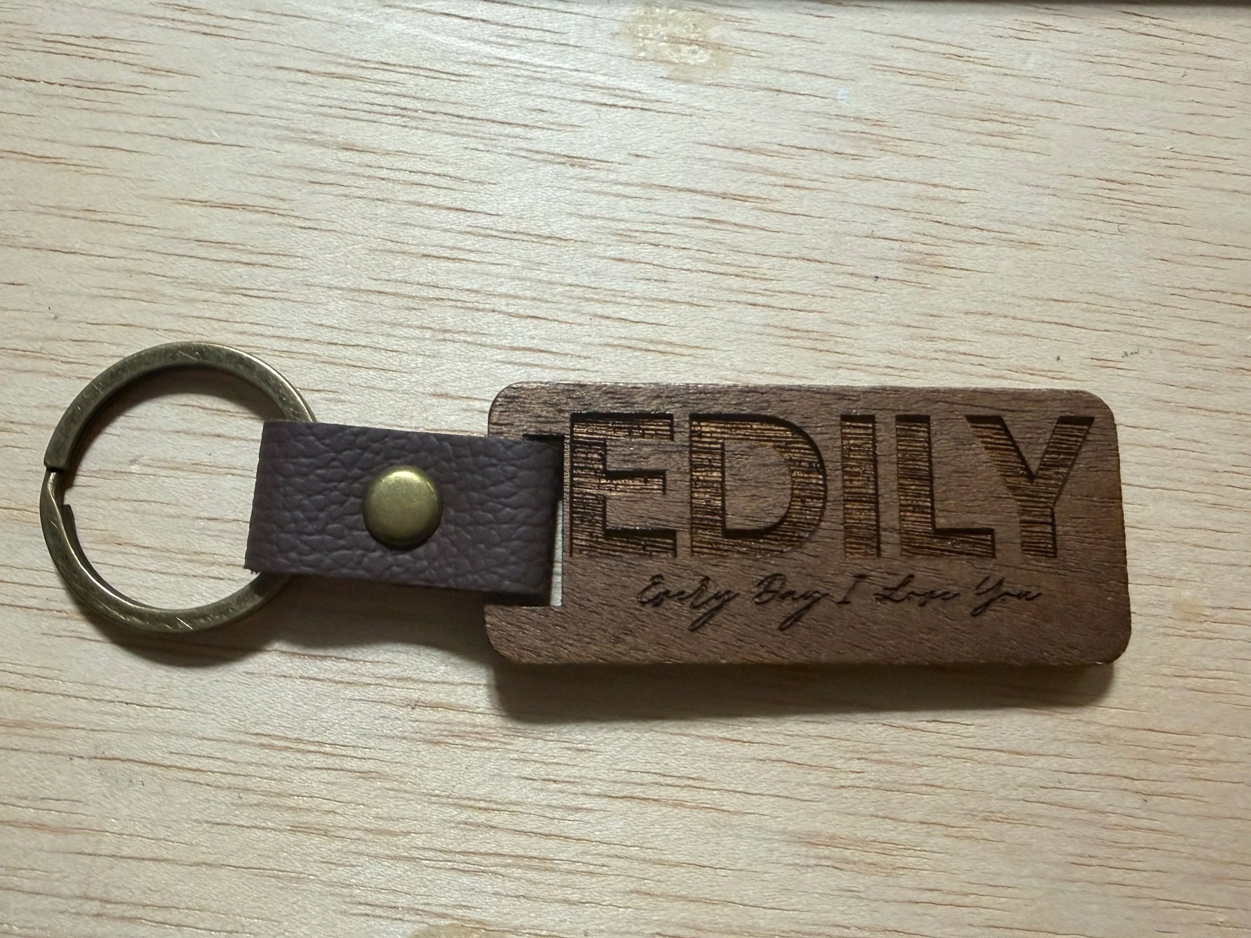 Close-up of a keychain with a leather strap and a wooden tag engraved with partially visible text, on a light wooden surface.