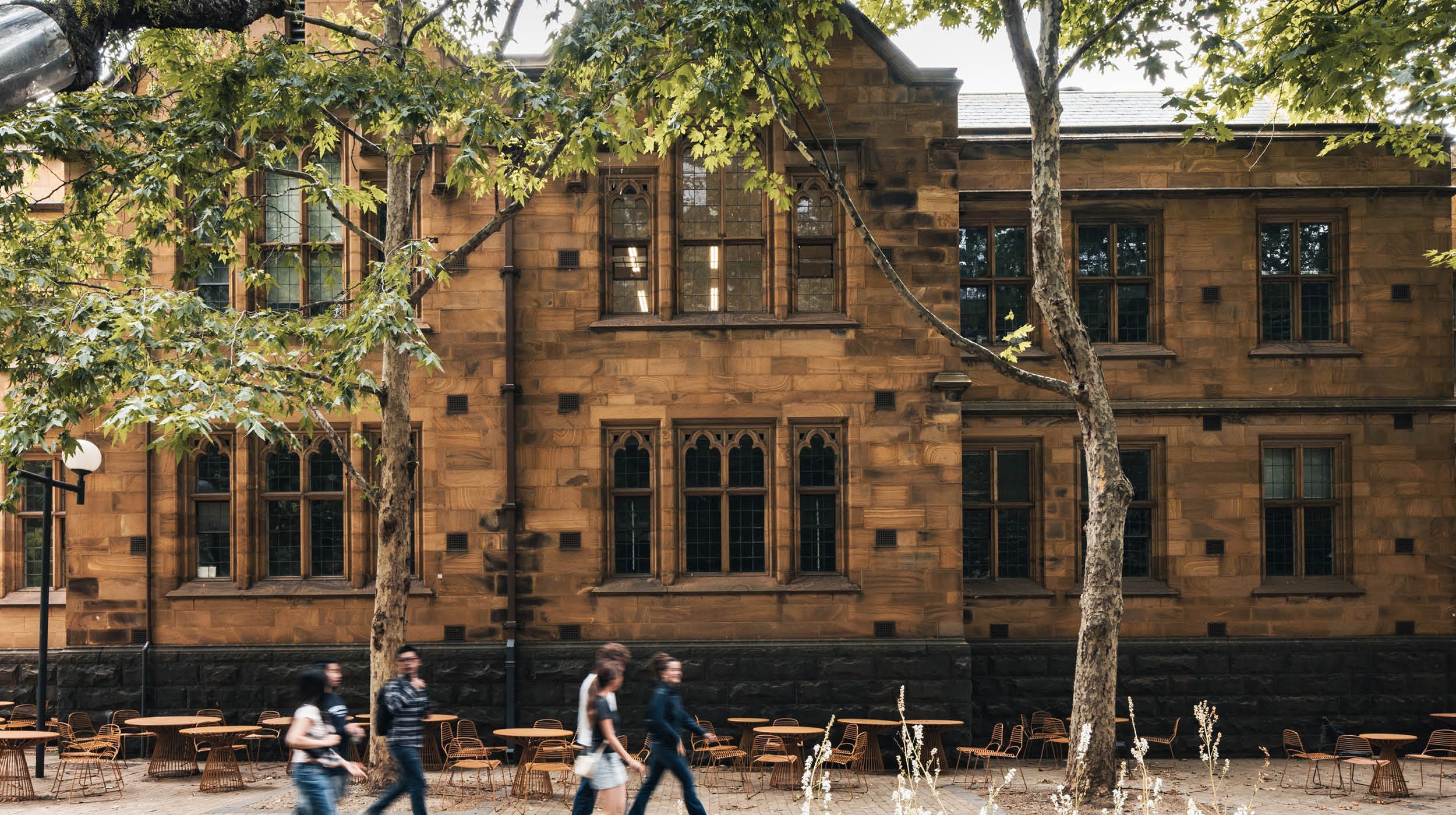 A historic brownstone building with gothic-style windows partially obscured by large green trees in an urban setting. There are outdoor tables and chairs at the sidewalk level, with people walking past.