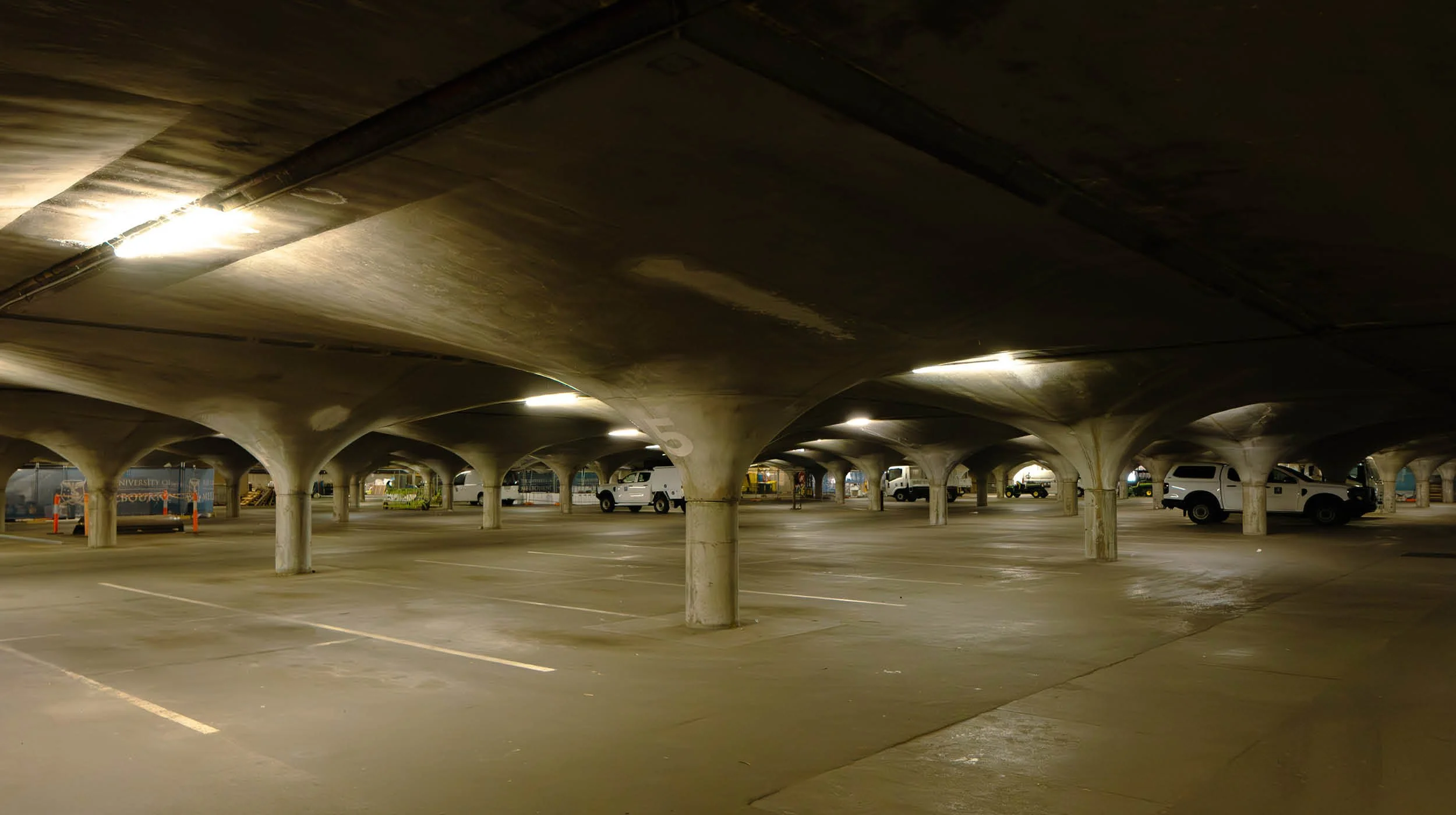 Underground parking garage with concrete ceiling supported by columns, dim lighting, and a few parked cars.