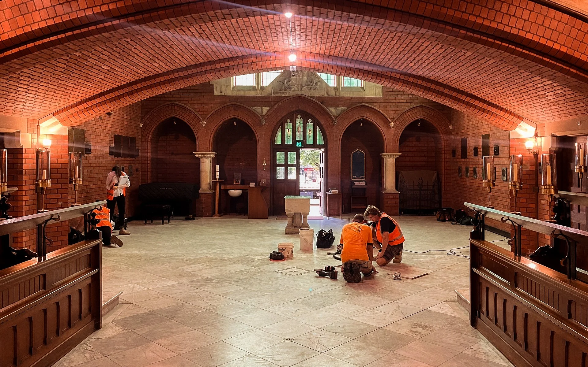 Interior of a church under renovation with workers installing and repairing flooring, with brick walls, windows, and religious statues visible in the background.