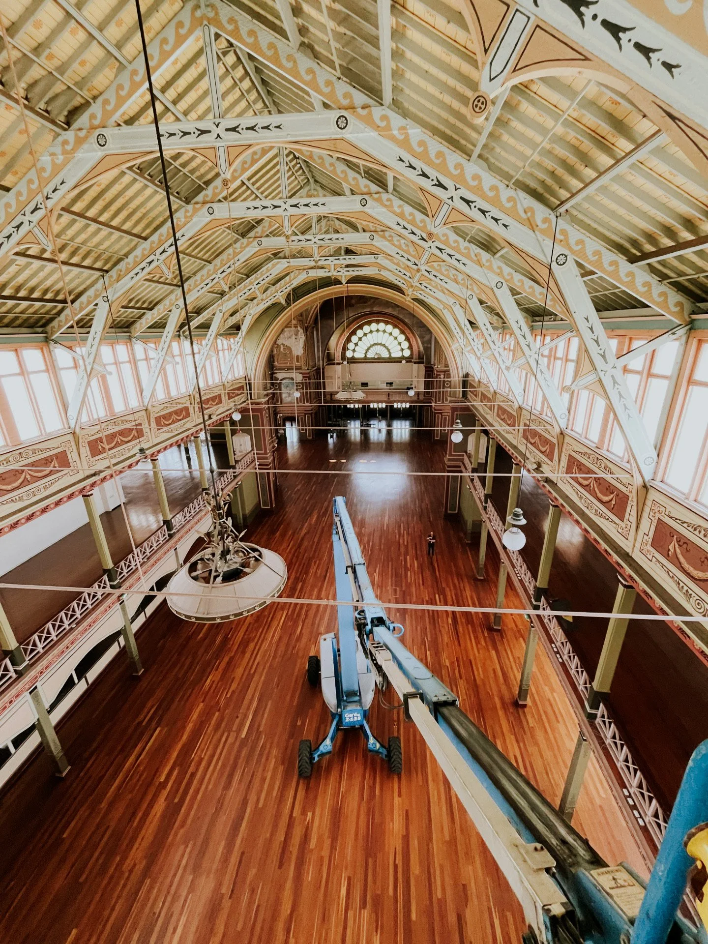 Interior of a historic building with ornate architectural details, high arched ceiling, and large windows. A blue boom lift is positioned in the center, indicating ongoing maintenance or restoration work.