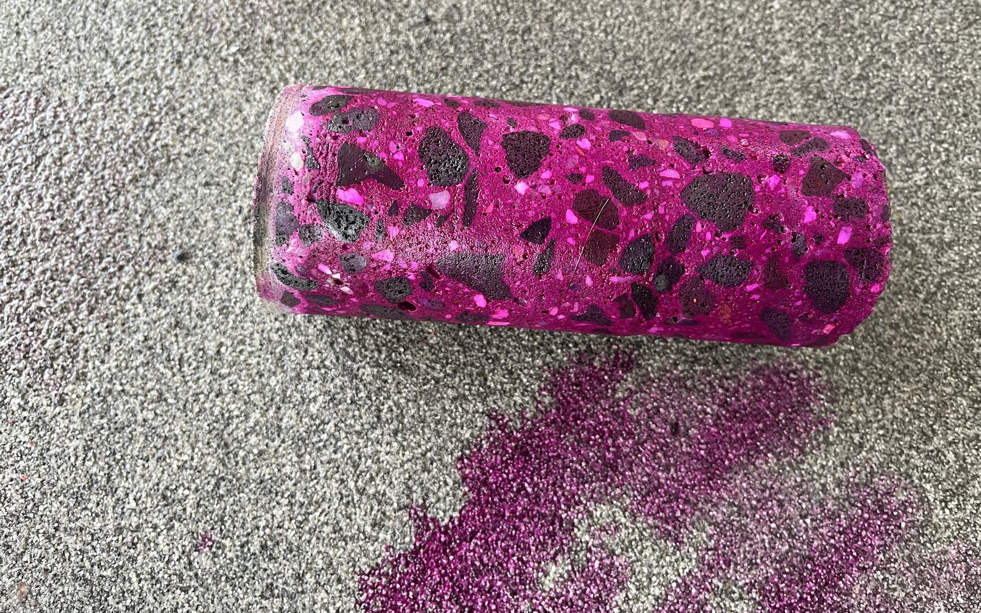 Close-up of a cylindrical grinder with pink and black speckled paint, spilled pink glitter below on a textured gray surface.