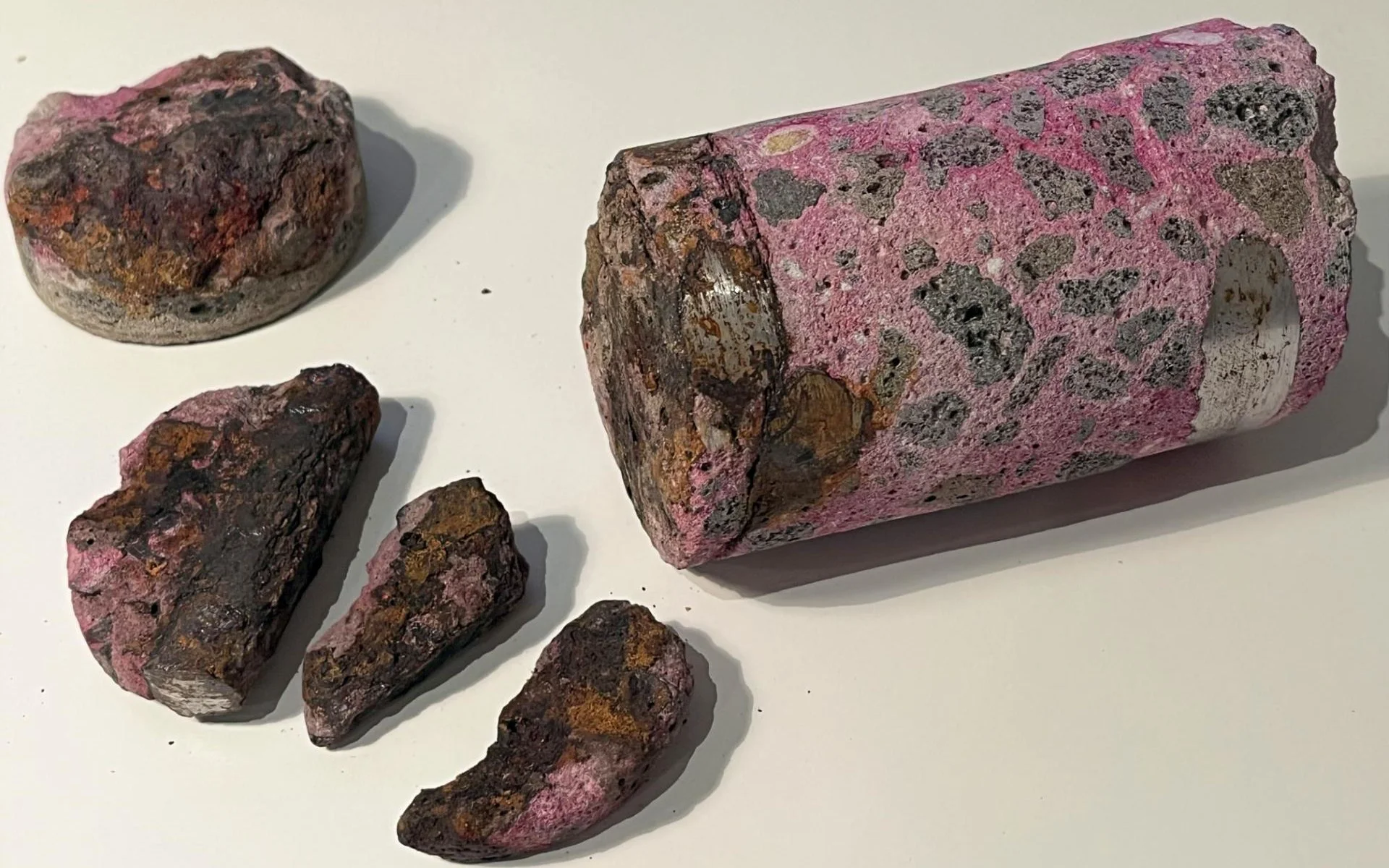 A collection of five igneous rocks with a pinkish hue and dark mineral inclusions, arranged on a white surface. Four smaller rocks are positioned around a larger, rectangular-shaped rock.