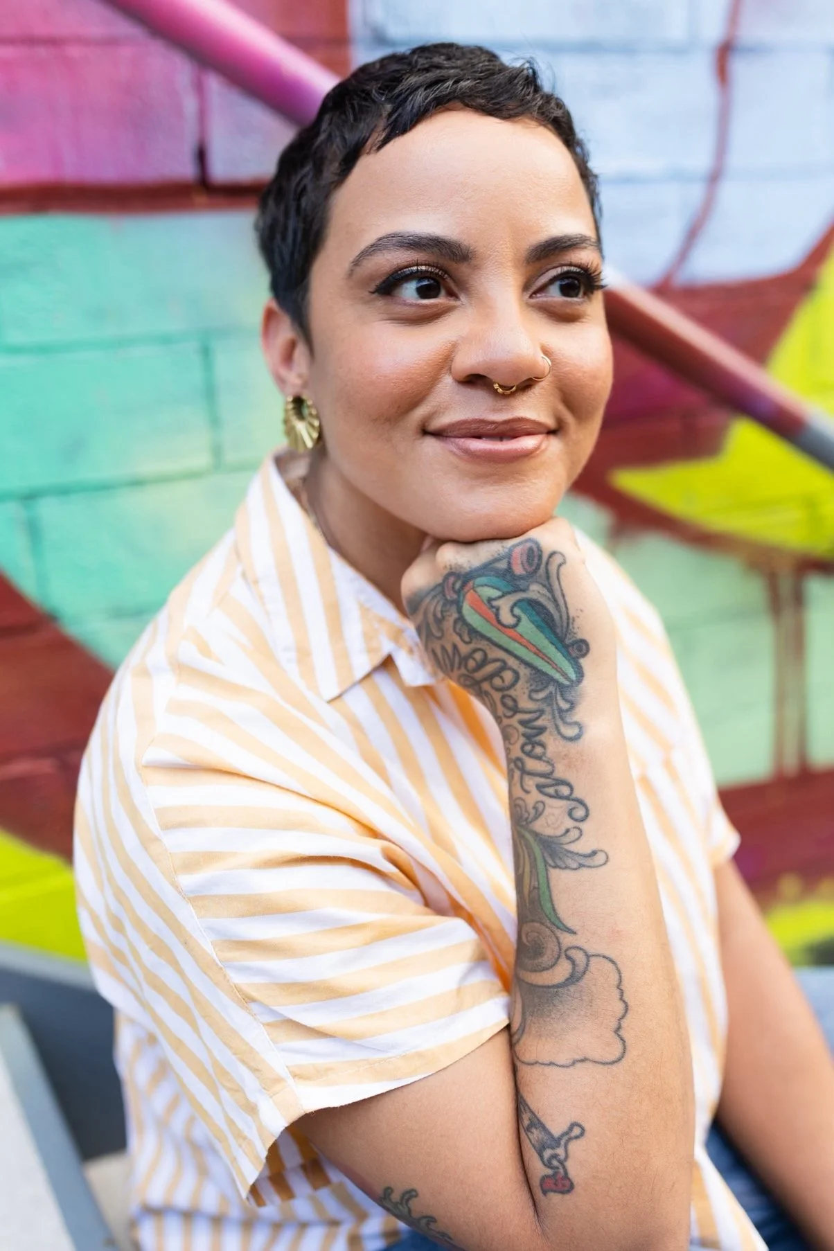 Portrait of voice actor Karlyn Granger, a Black woman with short dark hair and arm tattoos. She's wearing a yellow and white stripped shirt, smiling in front of a colorful mural.