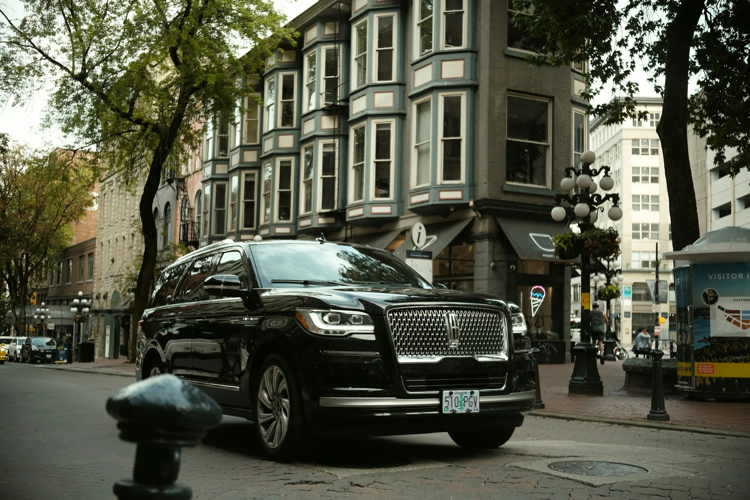 A city street scene featuring several parked cars, including a black Lincoln, in front of historic multi-story buildings with bay windows. There are trees lining the street, decorative street lamps, and signs indicating visitor information.