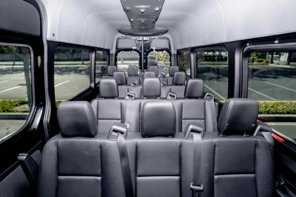 Interior of a passenger van with three rows of black leather seats, large windows on each side, and a view of an empty parking lot outside.