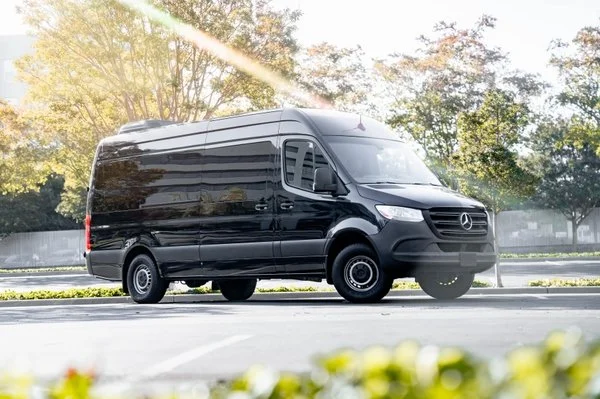 Black Mercedes-Benz Sprinter van parked on a street with trees and sunlight in the background.