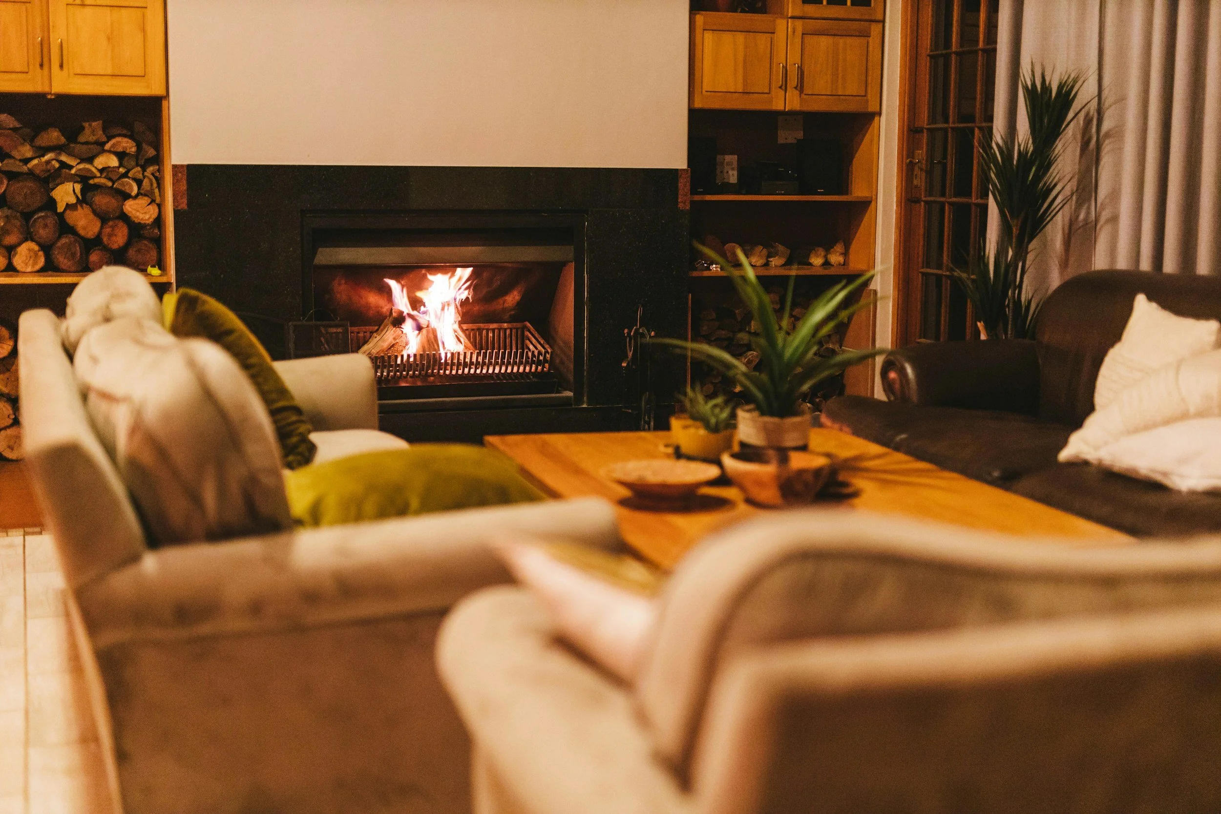 Living room with fireplace, wood logs, and furniture including couches and a wooden table with decorative bowls.
