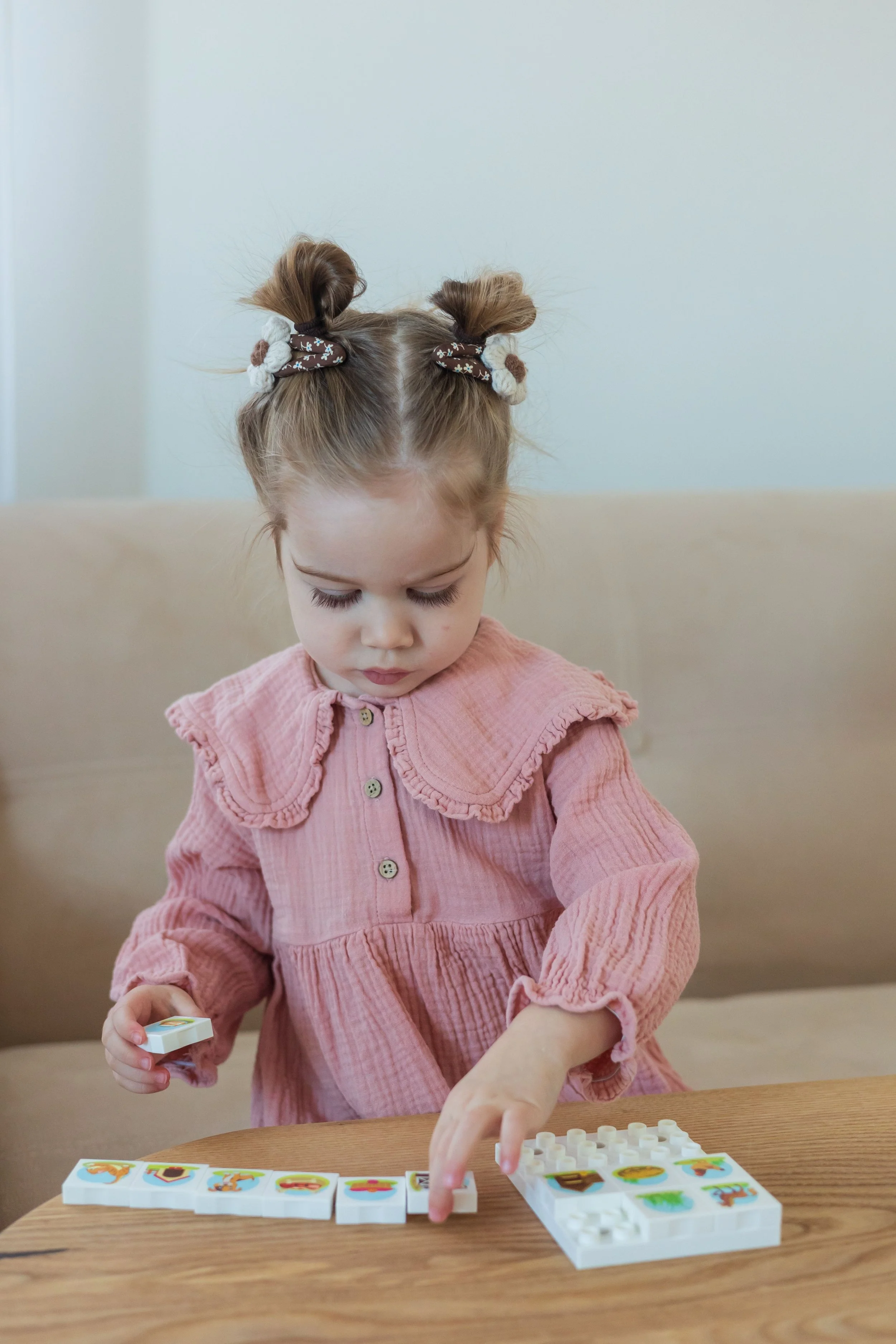 A young girl with brown hair in pigtails wearing a pink dress playing a tile-matching game at a wooden table.