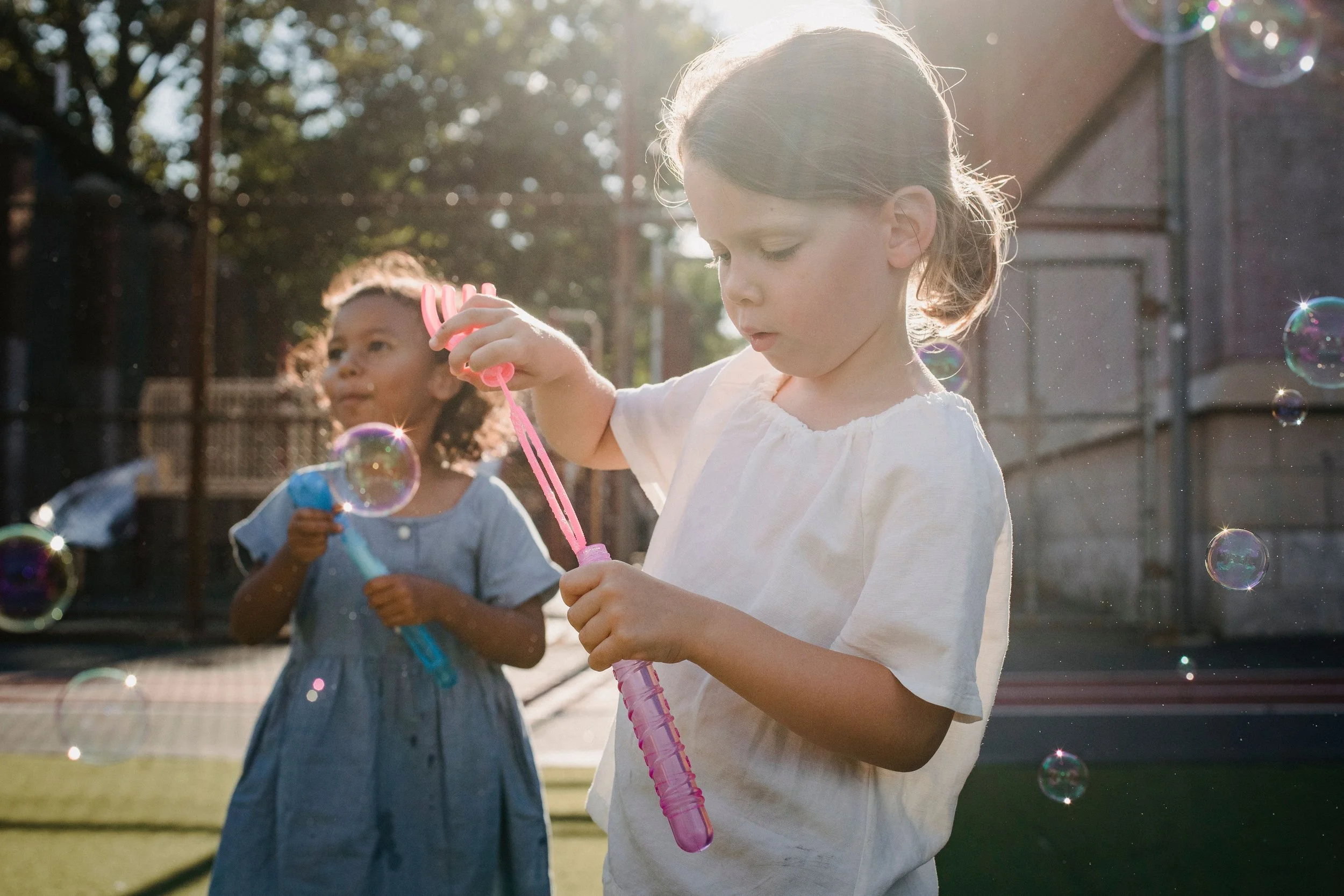 Two young girls playing with bubbles outdoors, with sunlight shining behind them and a park setting in the background.