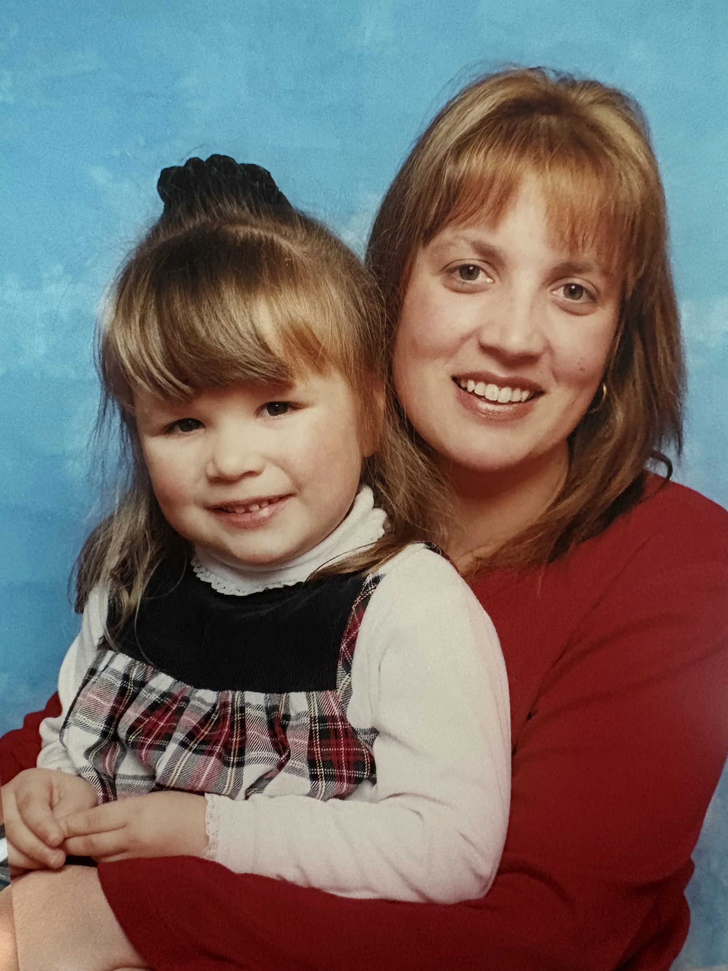 A woman with reddish-brown hair smiling, with a young girl with light brown hair and bangs, dressed in a white and plaid outfit, leaning in close to her against a blue background.