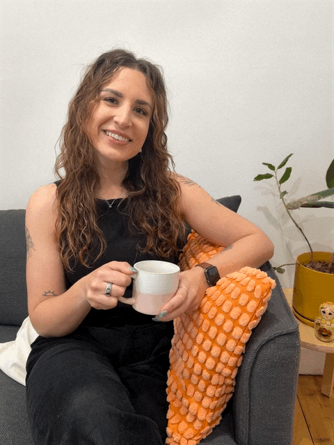 A young woman with long, curly hair sitting on a gray sofa, smiling and holding a white mug, with a potted plant and decorative items nearby.
