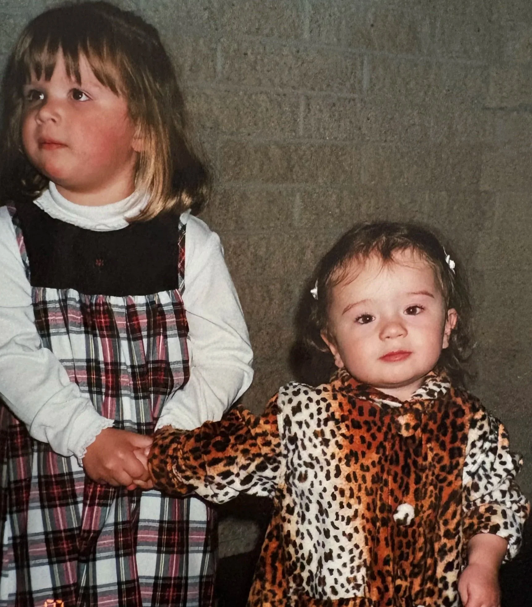 Close-up of two young girls with dark hair and light skin, one in a plaid dress and the other in a leopard print dress, standing against a gray brick wall, holding hands.