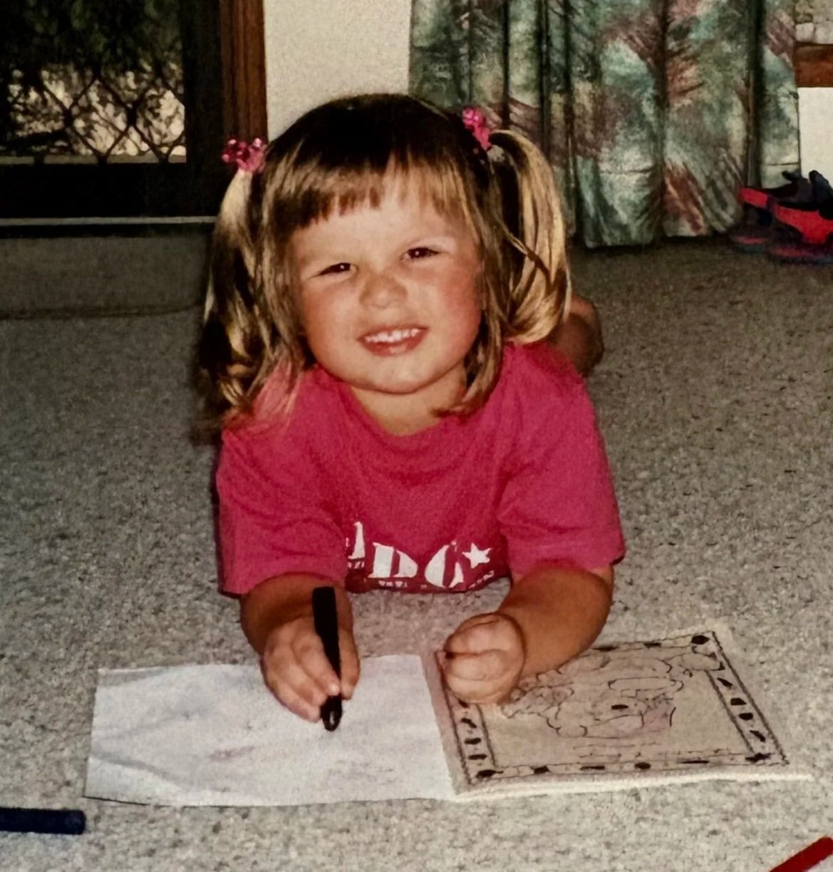 A young girl with blonde hair, tied in pigtails with pink hair ties, is sitting on a carpeted floor. She is smiling at the camera and coloring on a piece of paper with a black marker. She is wearing a pink T-shirt.