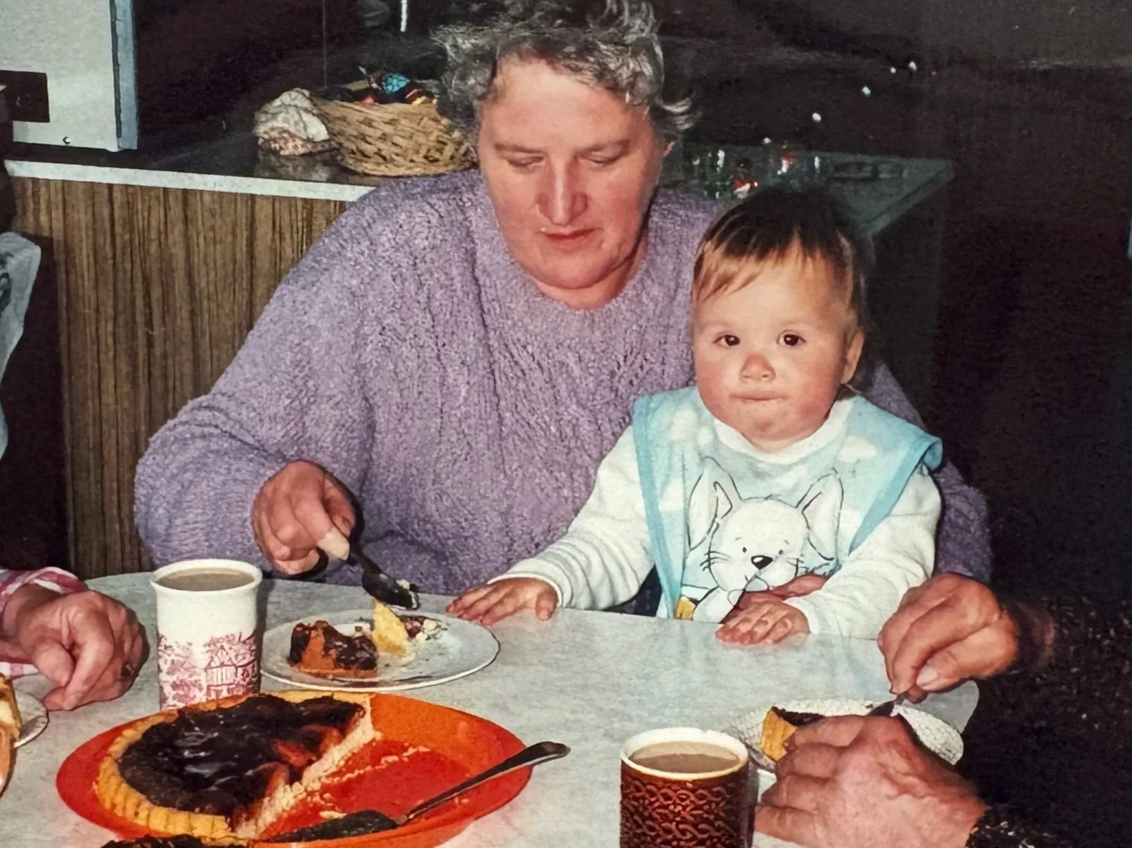 A woman and a young child sitting at a table with cake and coffee, sharing a meal together in a cozy, dimly lit indoor setting.