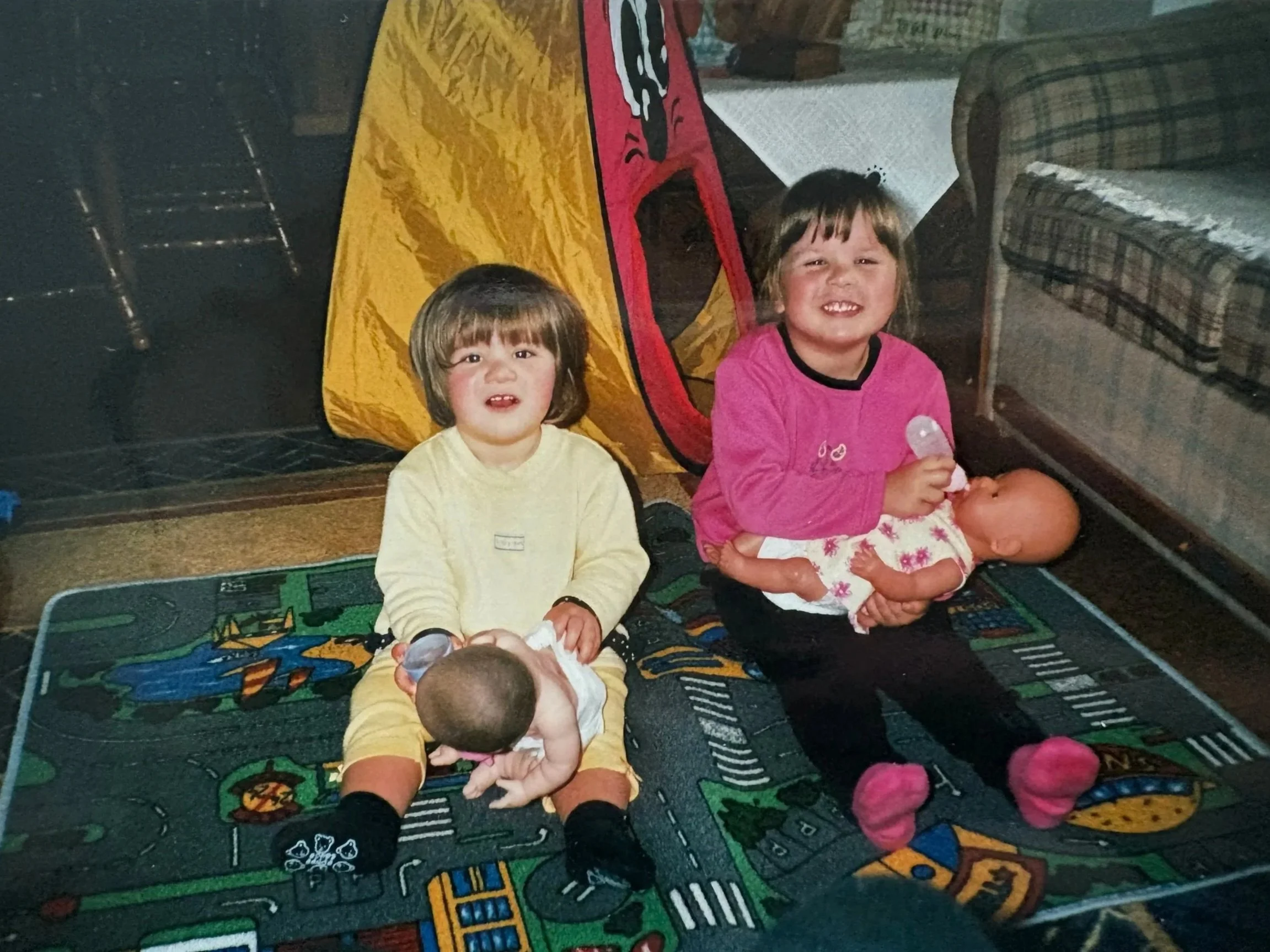 Two young children, a girl and a boy, sitting on a colorful play mat in a living room, with a yellow and red tent behind them and a baby lying on the girl’s lap. The girl is wearing a pink top and the boy a yellow shirt. They are smiling and holding toys.