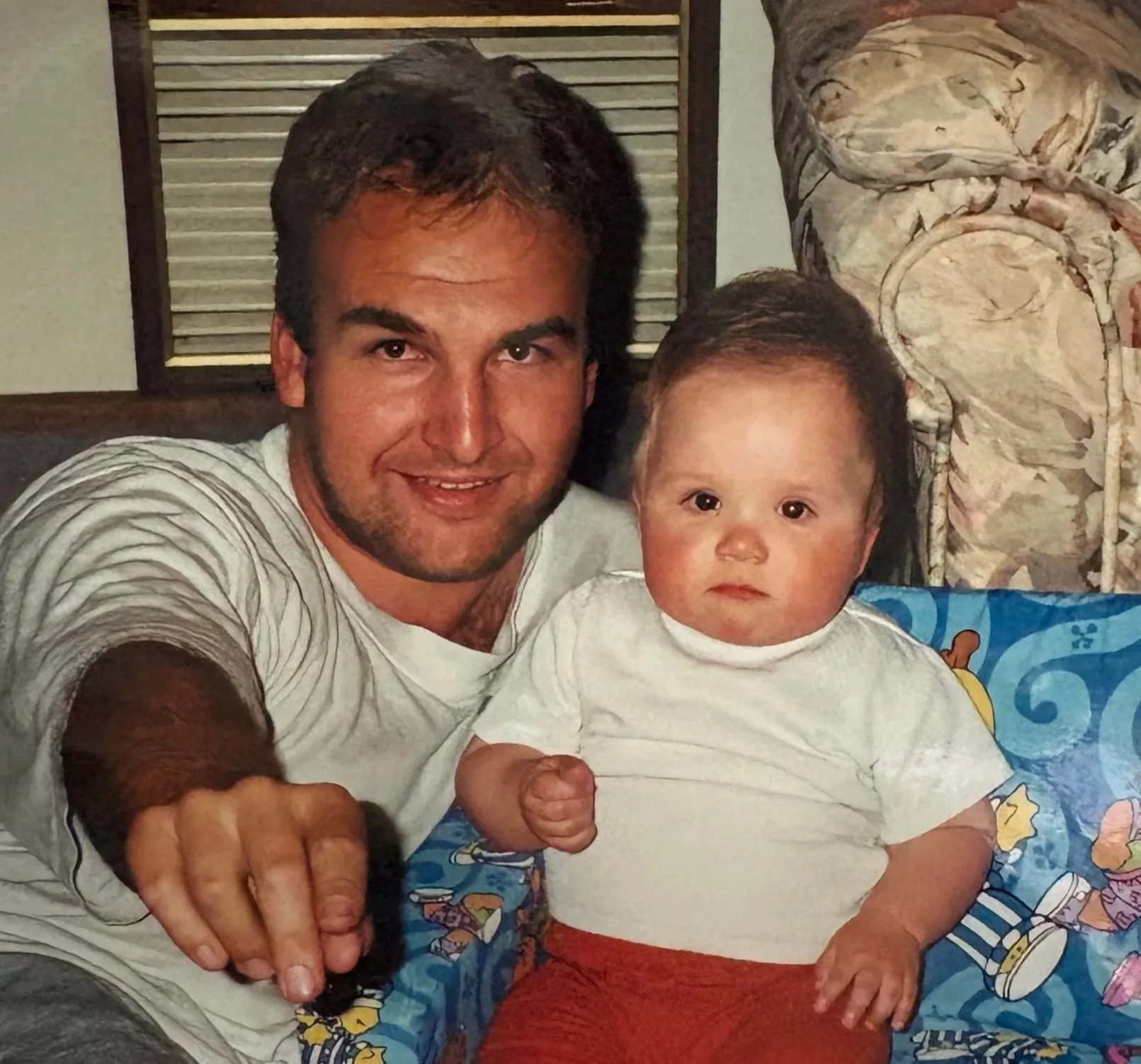 A man and a young boy sitting together, with the man grabbing the boy's hand. The man has dark hair, a beard, and is wearing a striped shirt. The boy has short dark hair, large eyes, and is wearing a white T-shirt and red shorts. They are sitting on a colorful blanket with cartoons, in a room with a wooden window in the background.