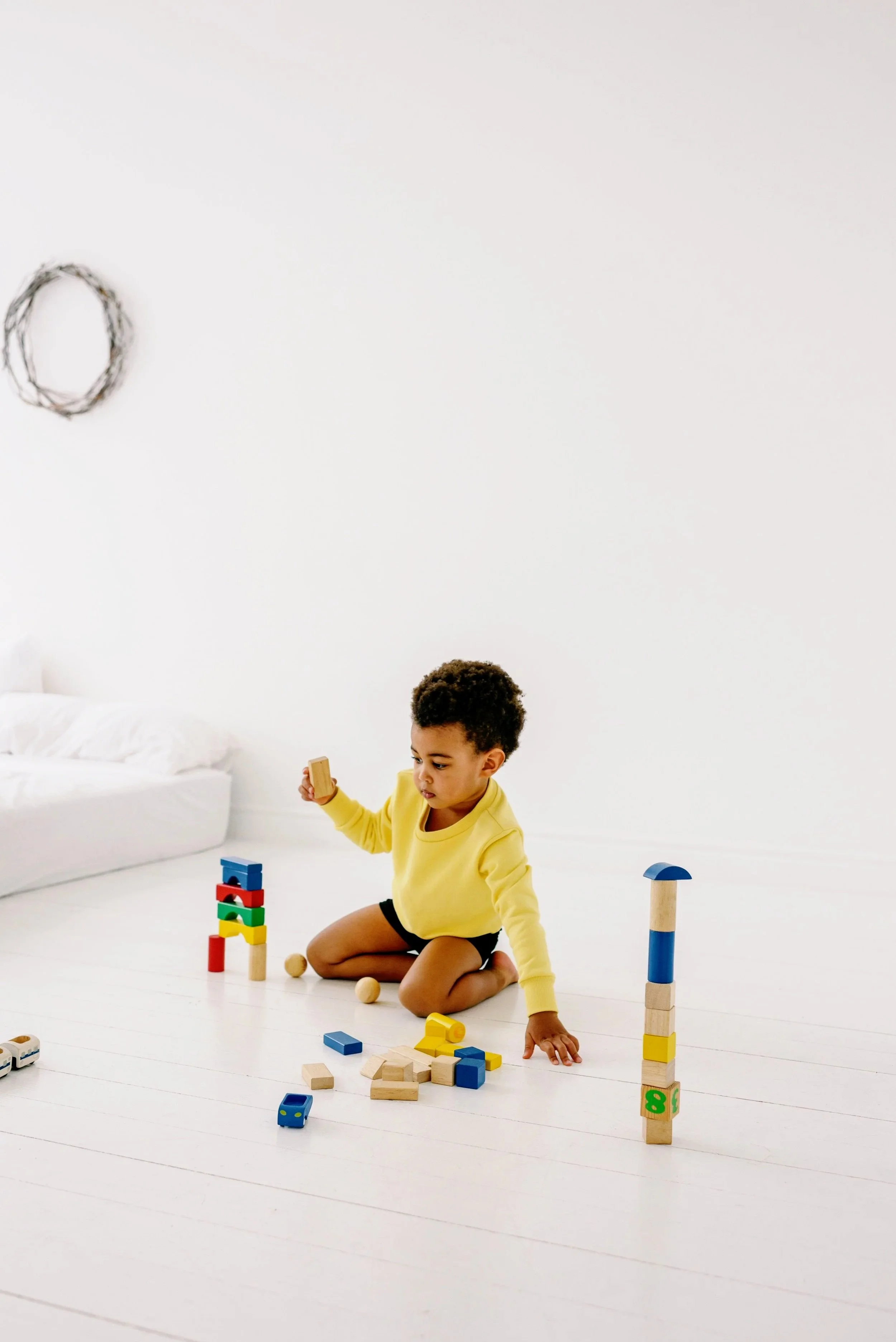 Young child playing with colorful wooden block stacking toys on a white floor in a bright, minimalist room.