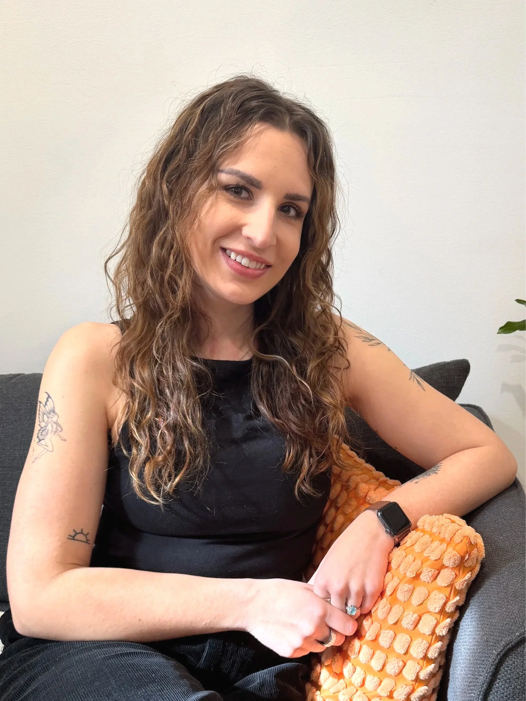 A woman with long wavy brown hair and tattoos on her arms sitting on a sofa smiling at the camera, with a plain white wall behind her.